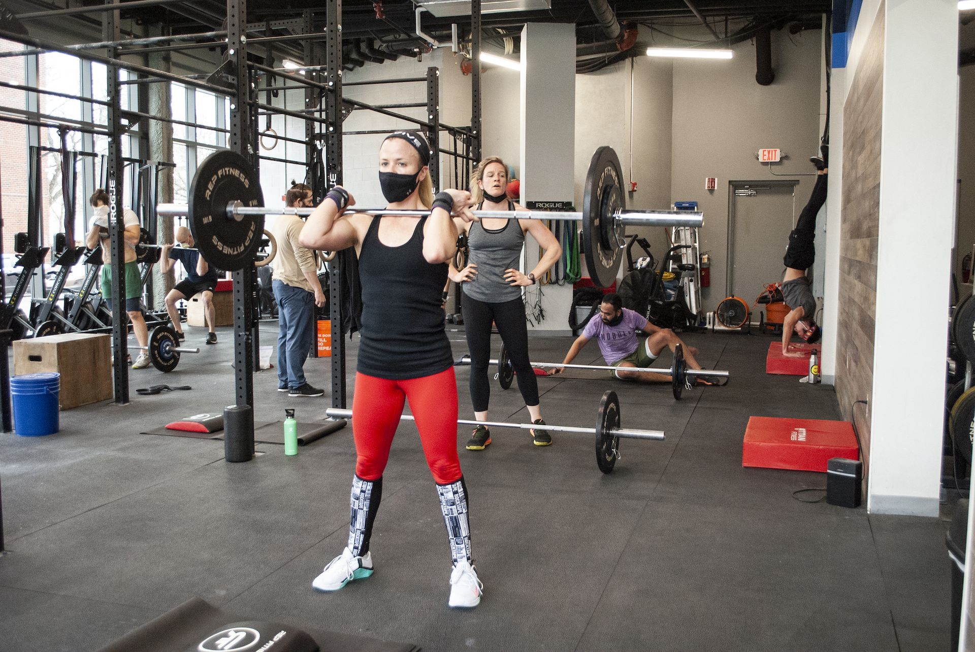Woman in mask lifts barbell in gym, others workout nearby.
