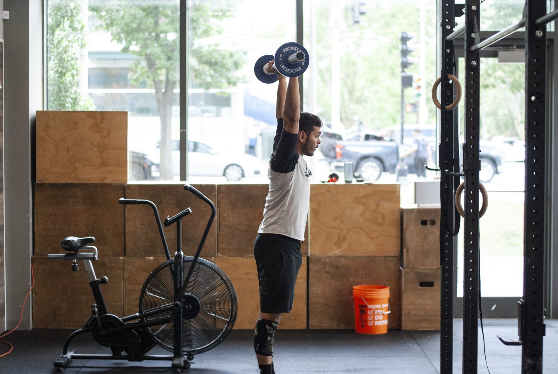 Man lifting barbell overhead in gym, wearing black shorts, knee brace.