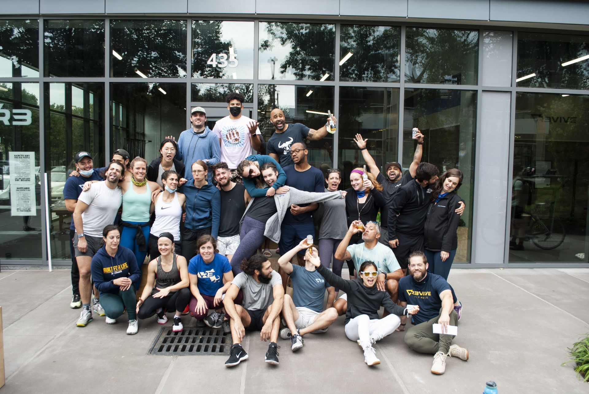 Group of people pose outside building with glass windows. Some crouch, some lift person. All smiling.