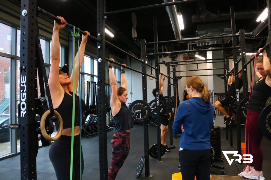 People doing pull-ups in a gym. Women with black tops and leggings. One using a resistance band.