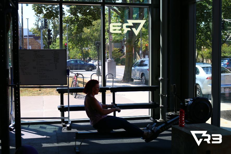 Woman rowing indoors, seen through a window. Sunlight streams in.