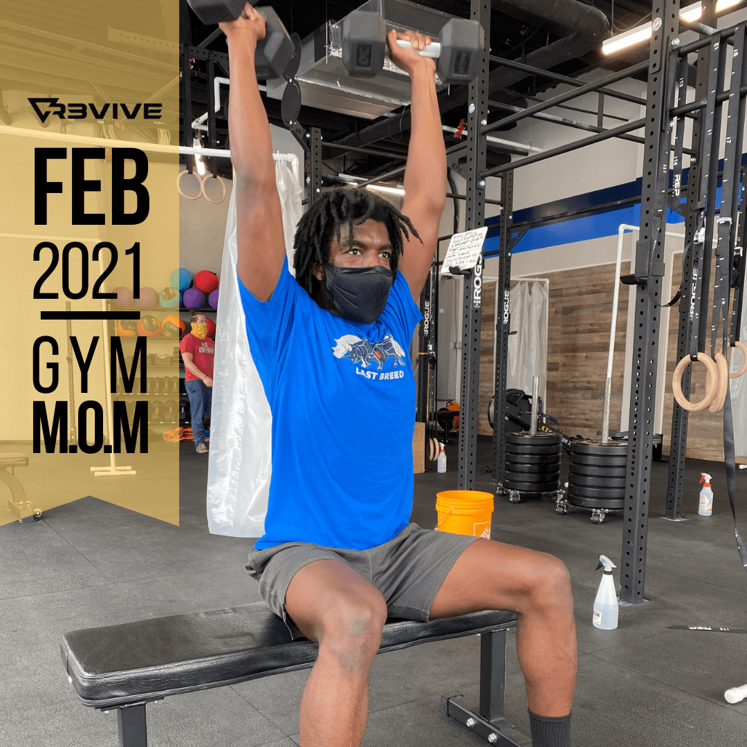 Man in blue shirt lifting dumbbells overhead while seated on a bench at a gym.