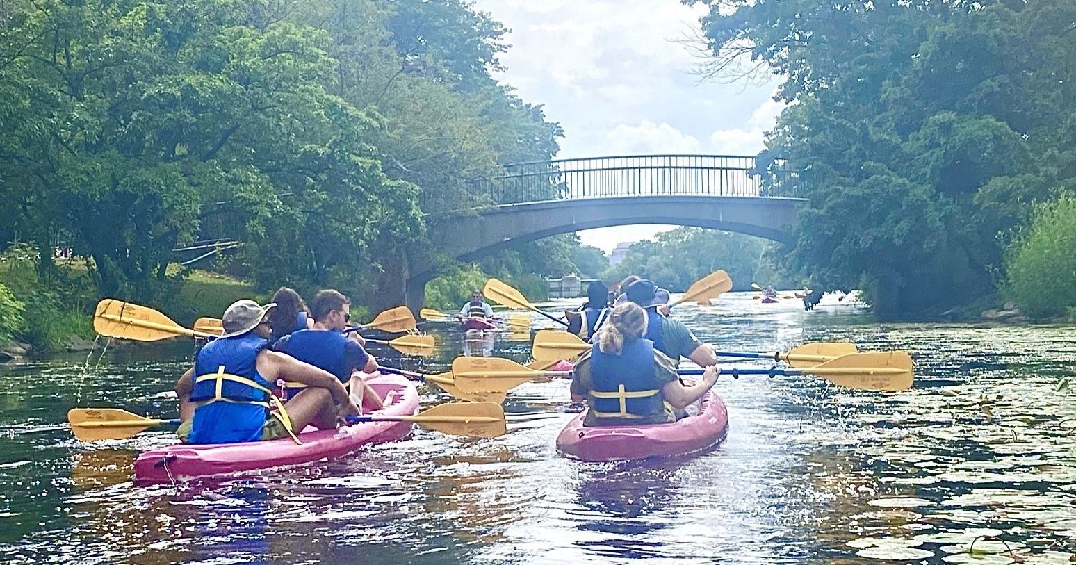 Kayakers paddle on a river under a stone bridge, surrounded by trees.