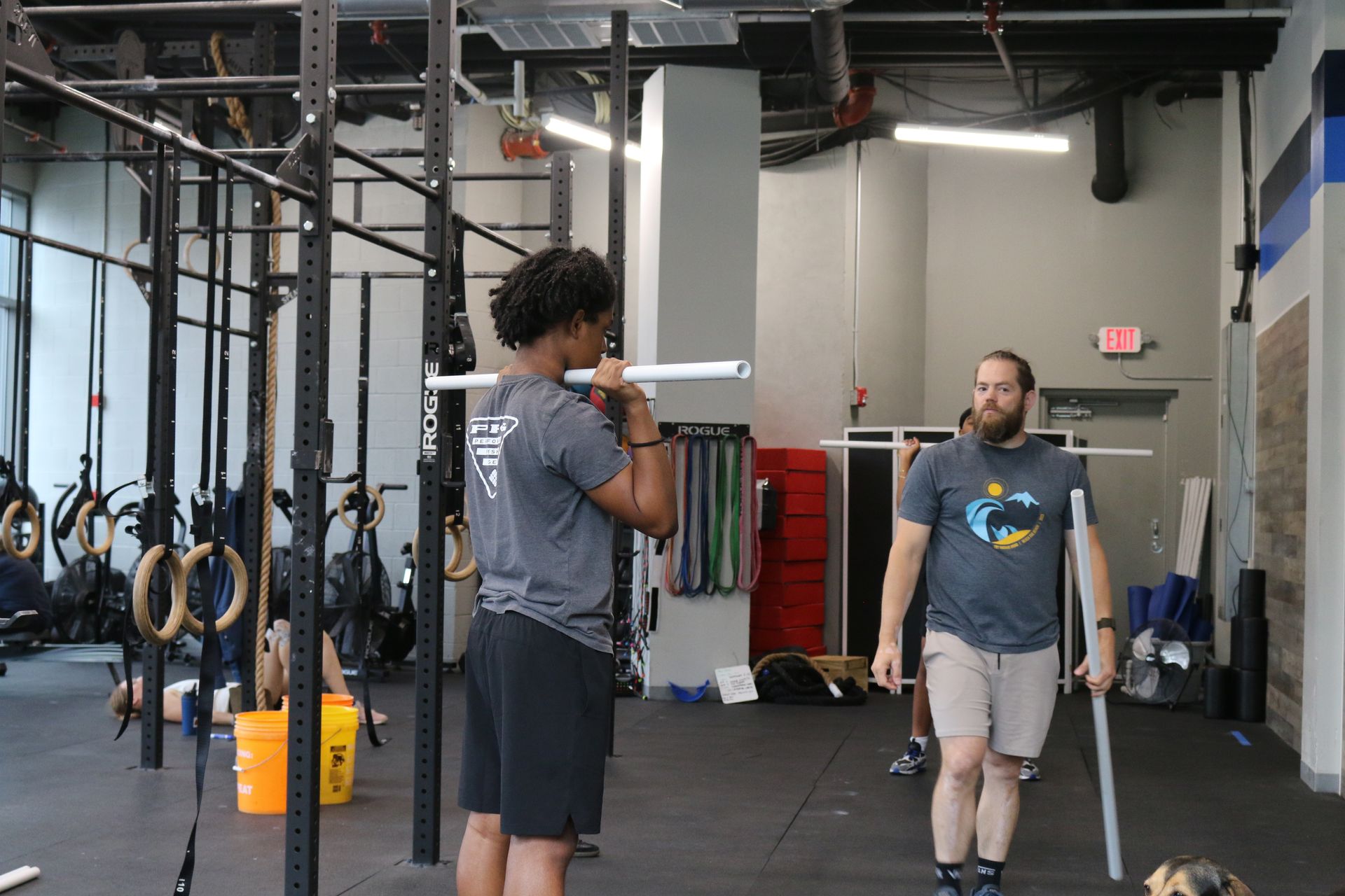 Man practicing overhead squat with a PVC pipe, instructor watches in a gym.
