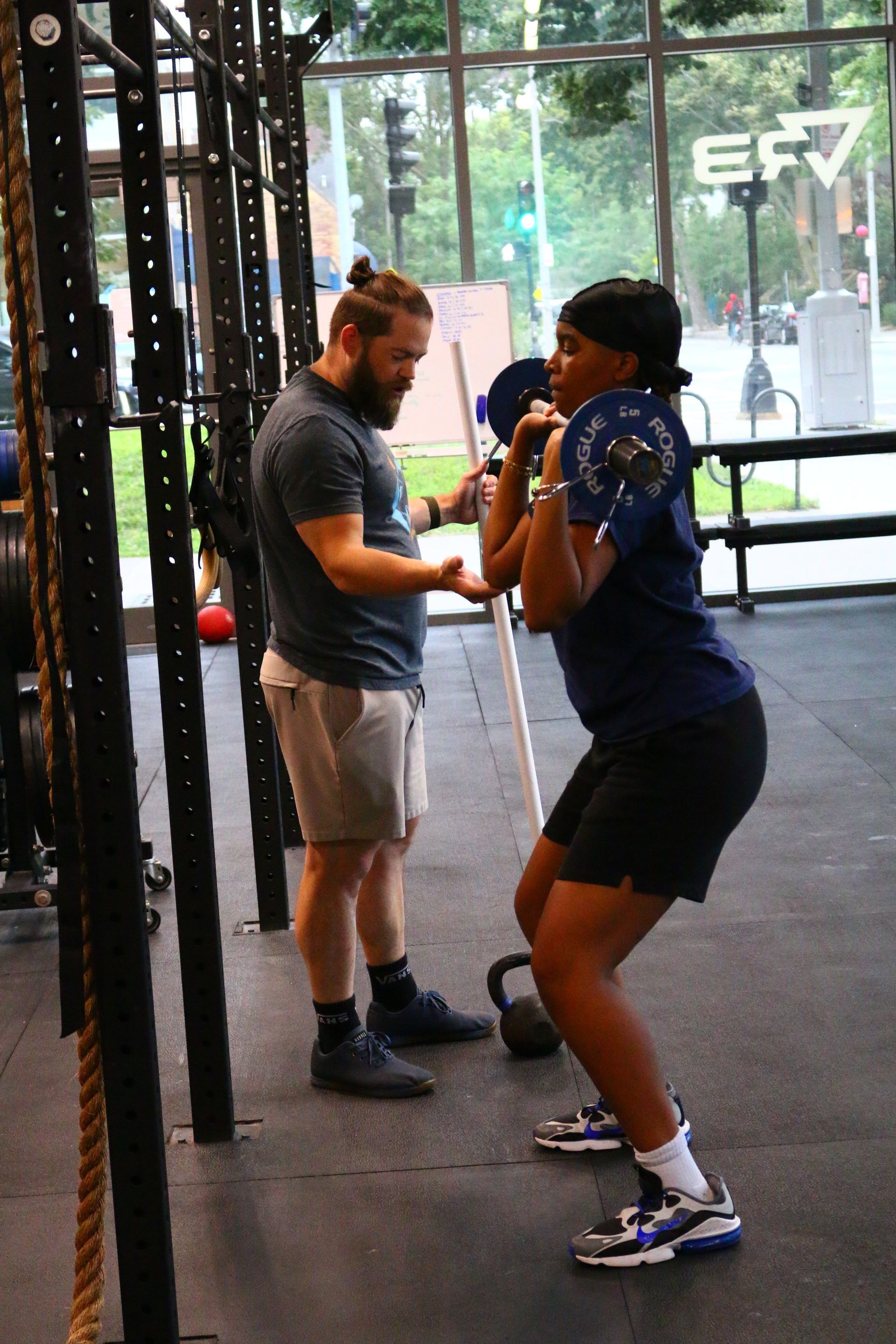 A woman performs a barbell squat while a coach observes in a gym.