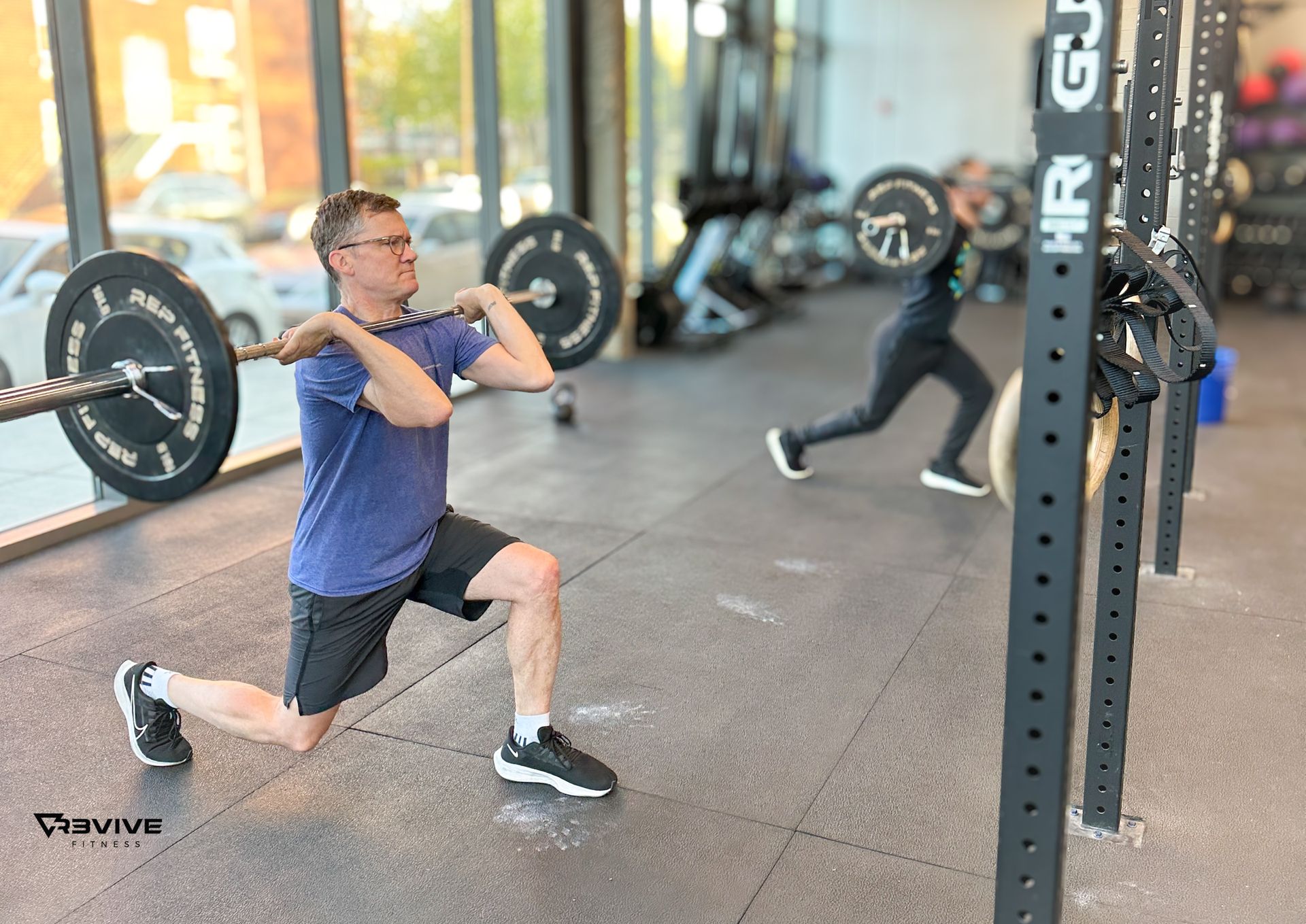 Man lunging with barbell at a gym; another person works out in the background.