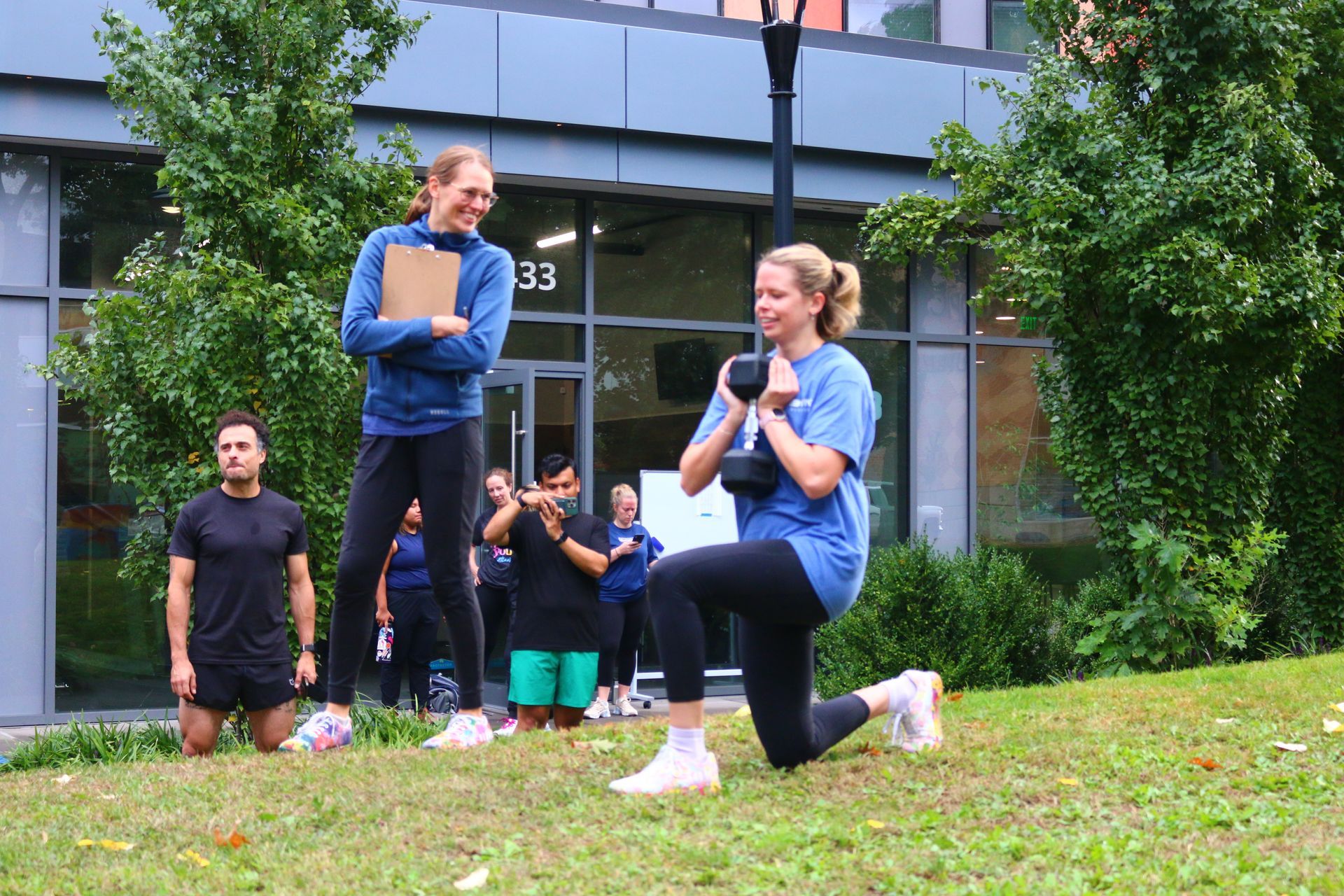 Woman doing lunges with a dumbbell, trainer watching. Outdoor workout in front of a building.