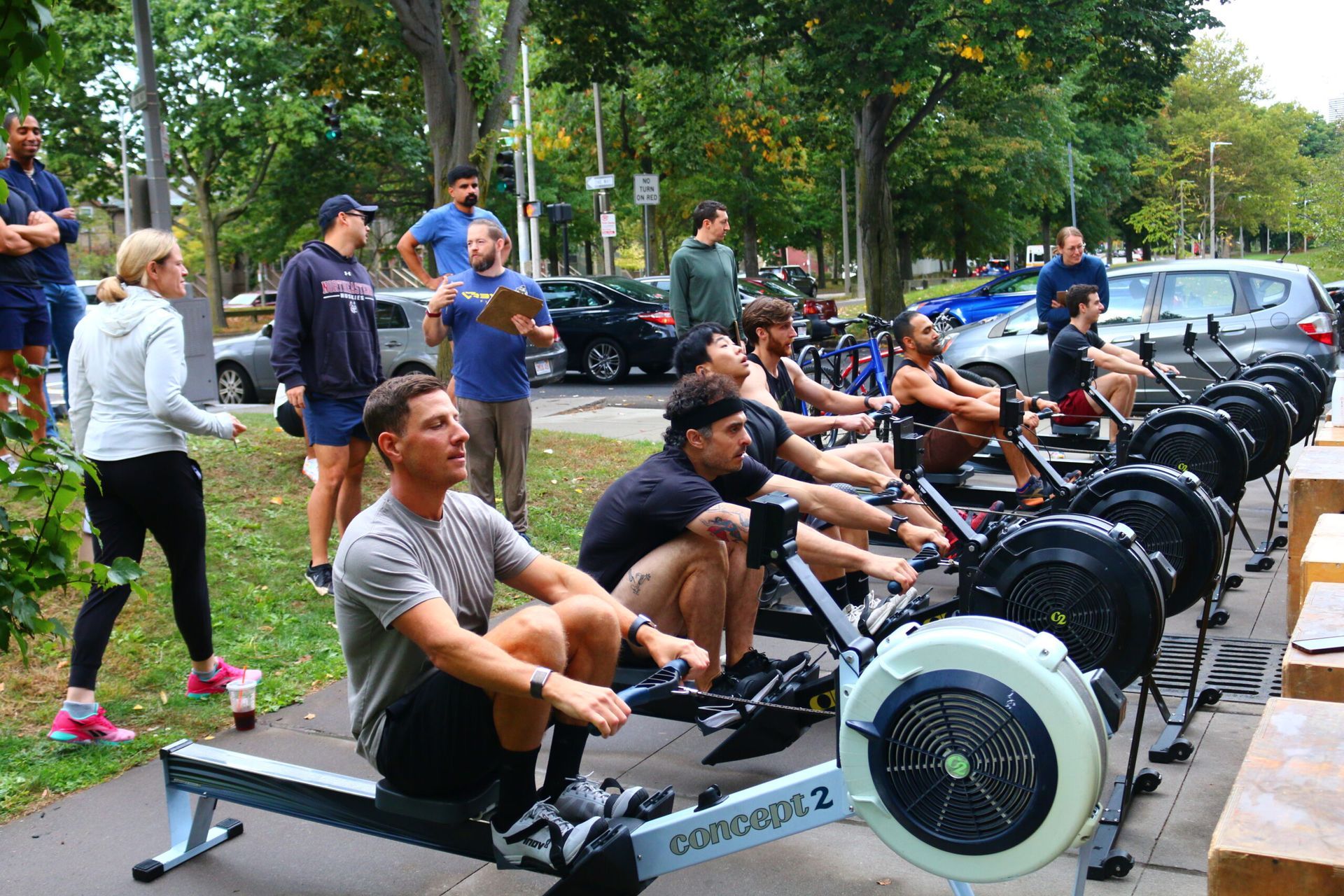 Rowers on machines outdoors, people observing; park setting.