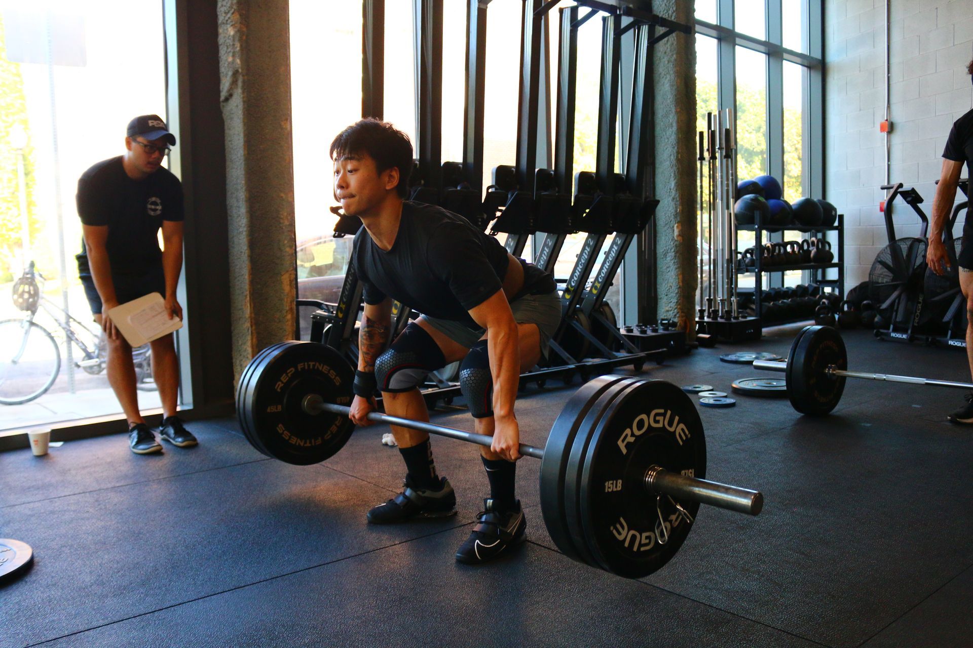 Man lifting barbell in a gym, wearing knee sleeves and black attire. Trainer watches.