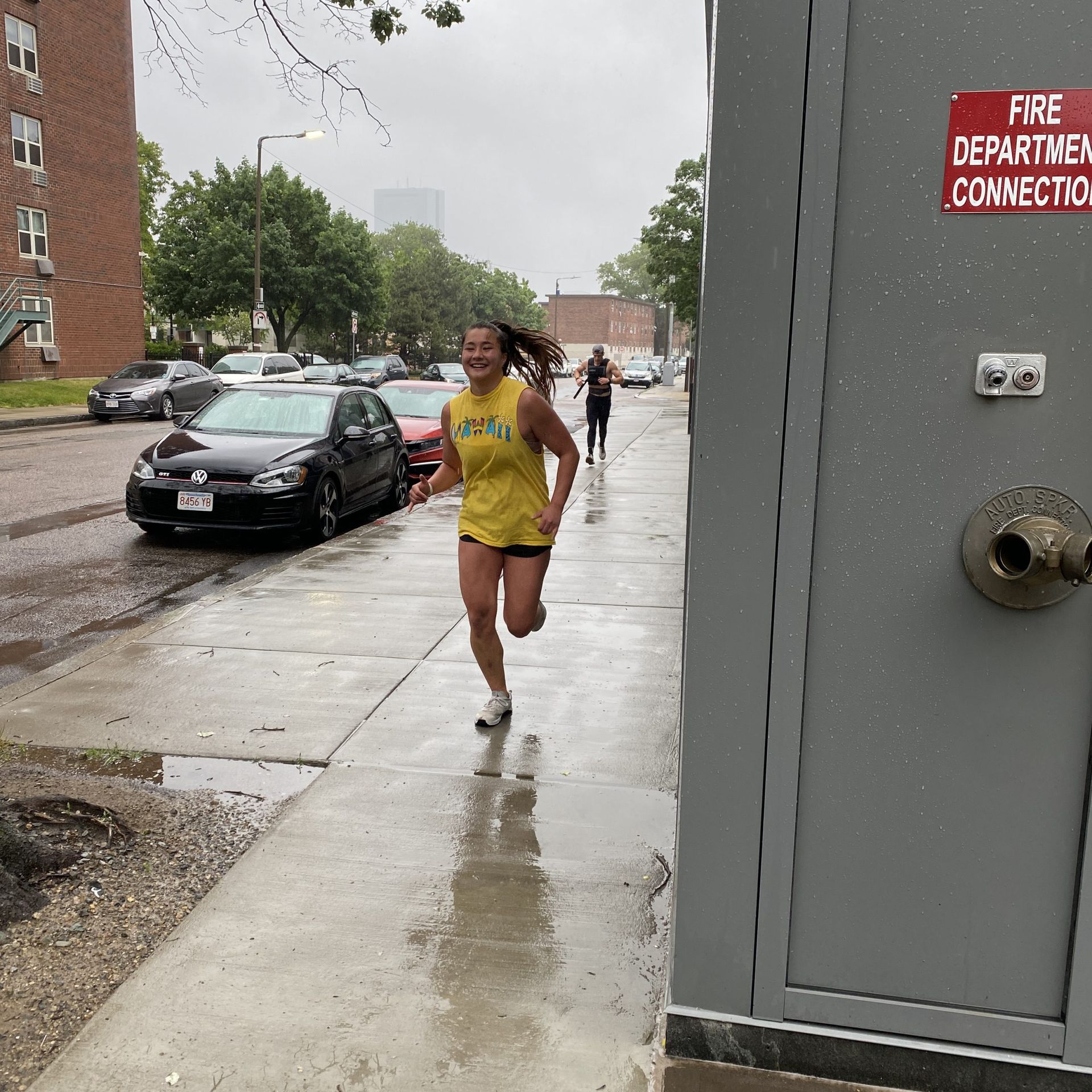 Woman runs happily down a wet sidewalk in the rain; cars and buildings in background.