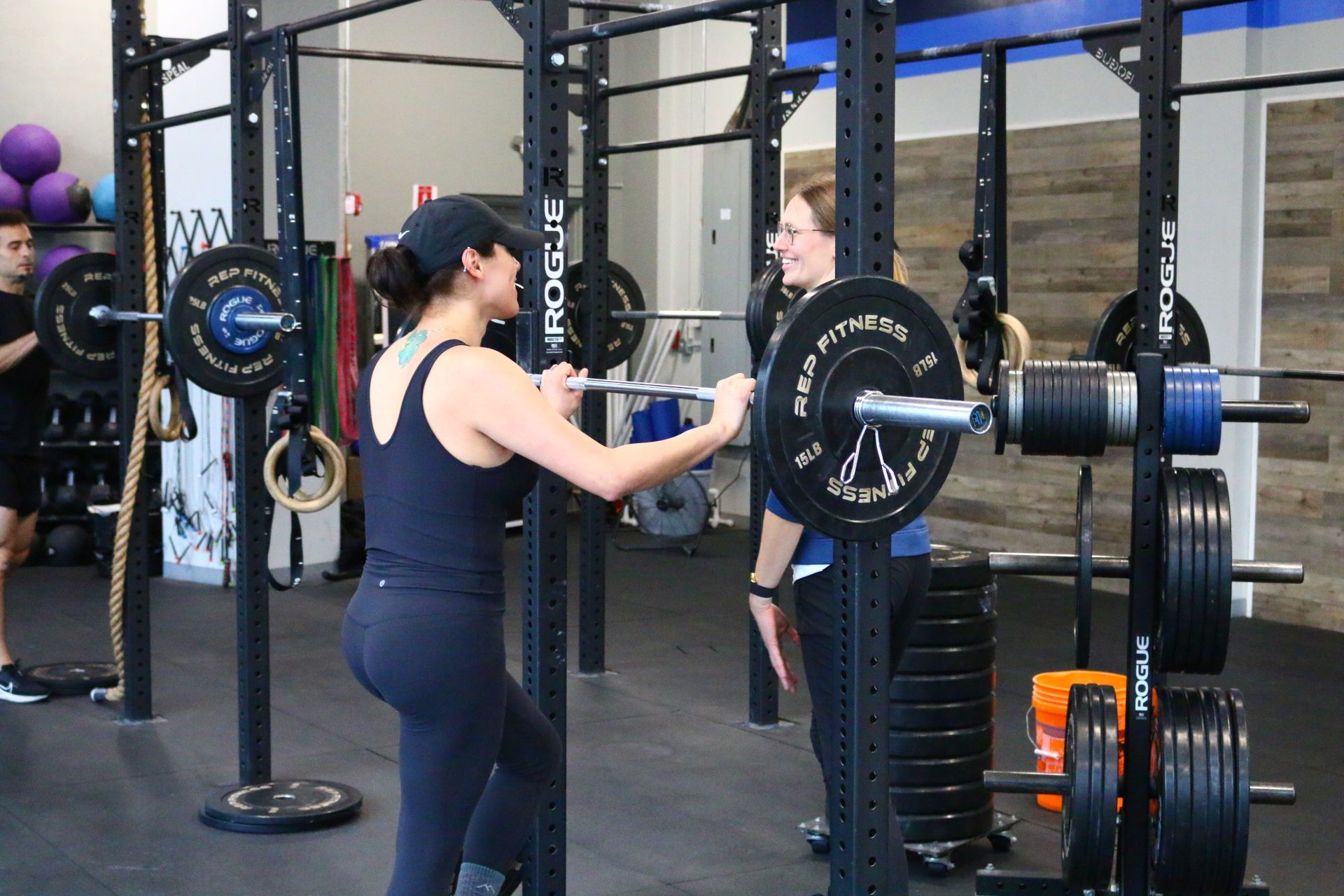 Woman preparing to lift a barbell with assistance from another woman in a gym setting.