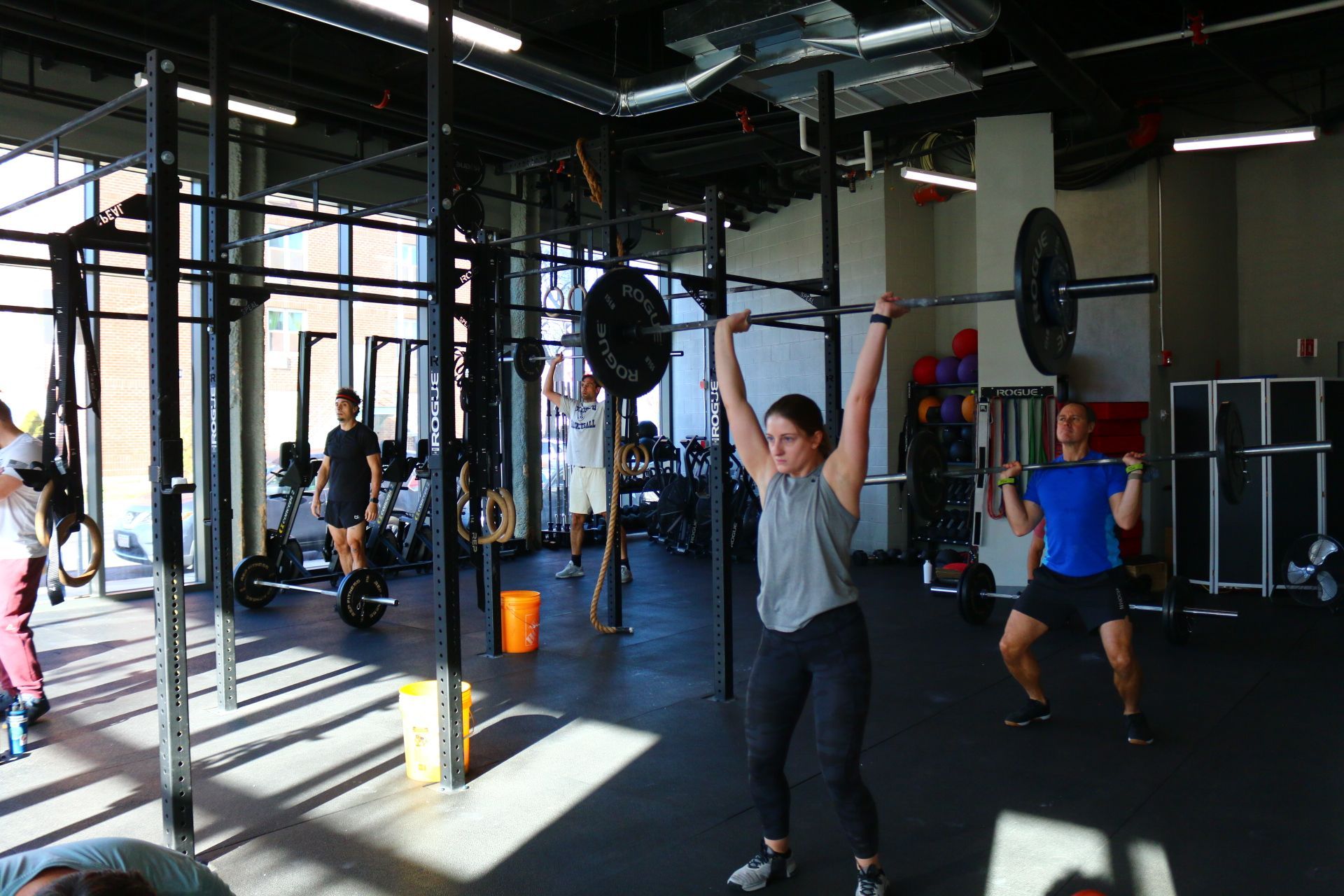 People working out in a gym, lifting weights. A woman overhead presses a barbell.