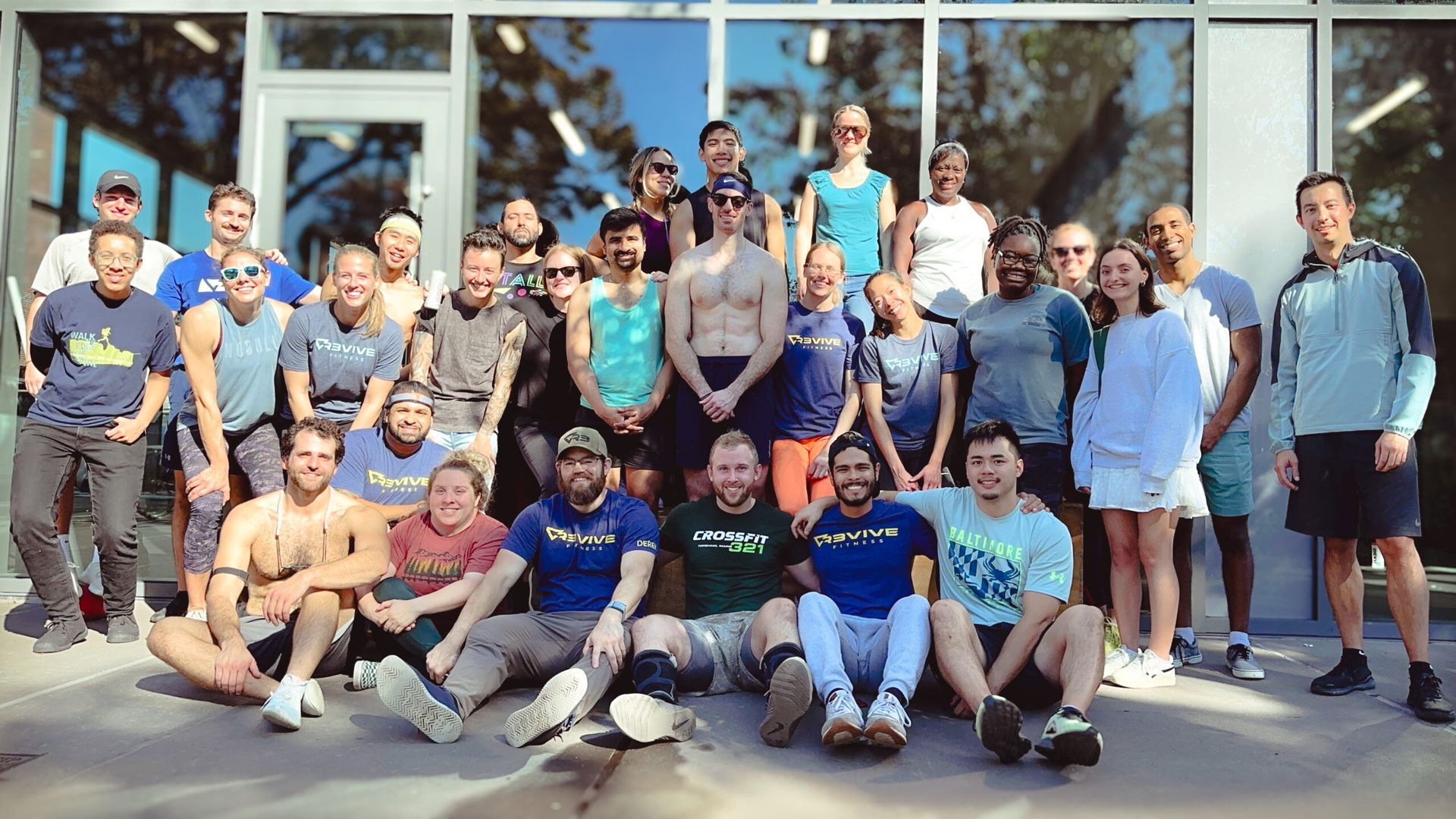 Group of diverse people smiling outside a building. Some are seated, others standing. Sunny, daytime.