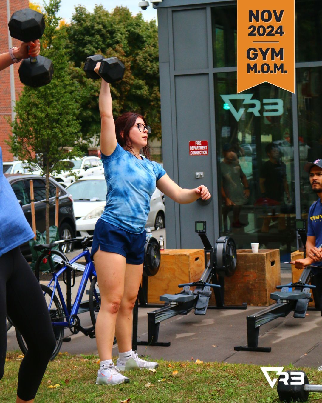 Woman lifting dumbbell overhead at an outdoor gym. Blue tie-dye shirt, dark shorts, white sneakers. NOV 2024 Gym M.O.M.
