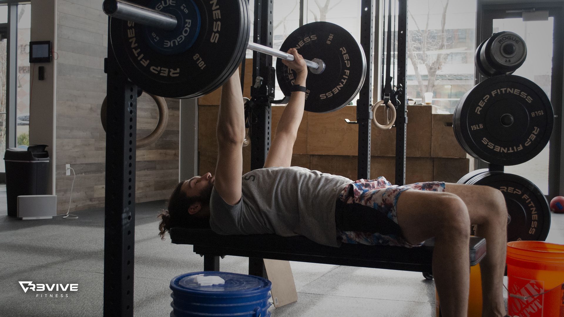 Man bench presses a barbell, lying on a black bench in a gym.