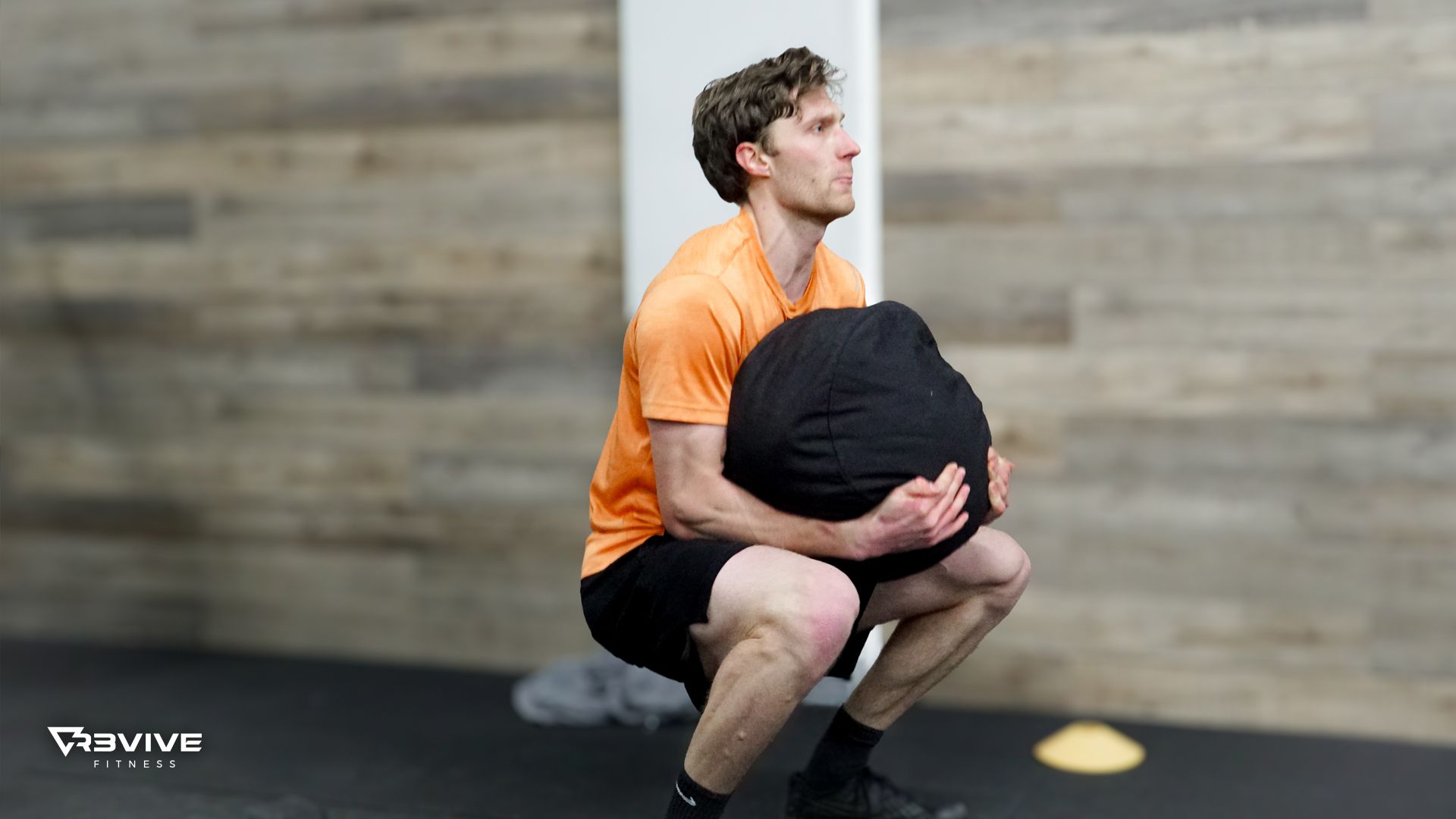 Man in orange shirt squats holding a black medicine ball. He's in a gym with wood paneling.