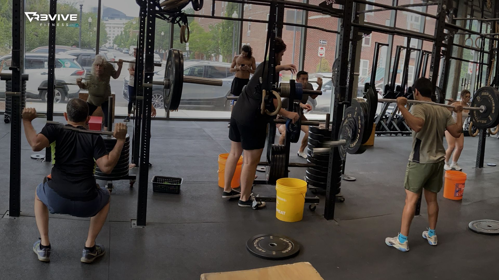 People are squatting with barbells in a gym with large windows. Some are loading weights.