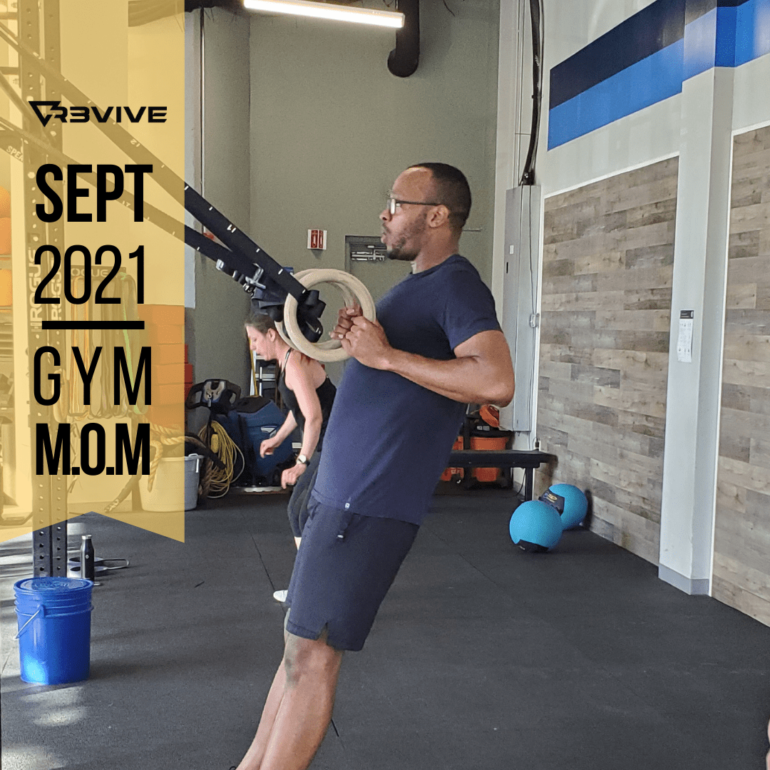 Man doing a ring row exercise in a gym. He's wearing a navy shirt and shorts. Blue bucket, rings, and a woman are visible.
