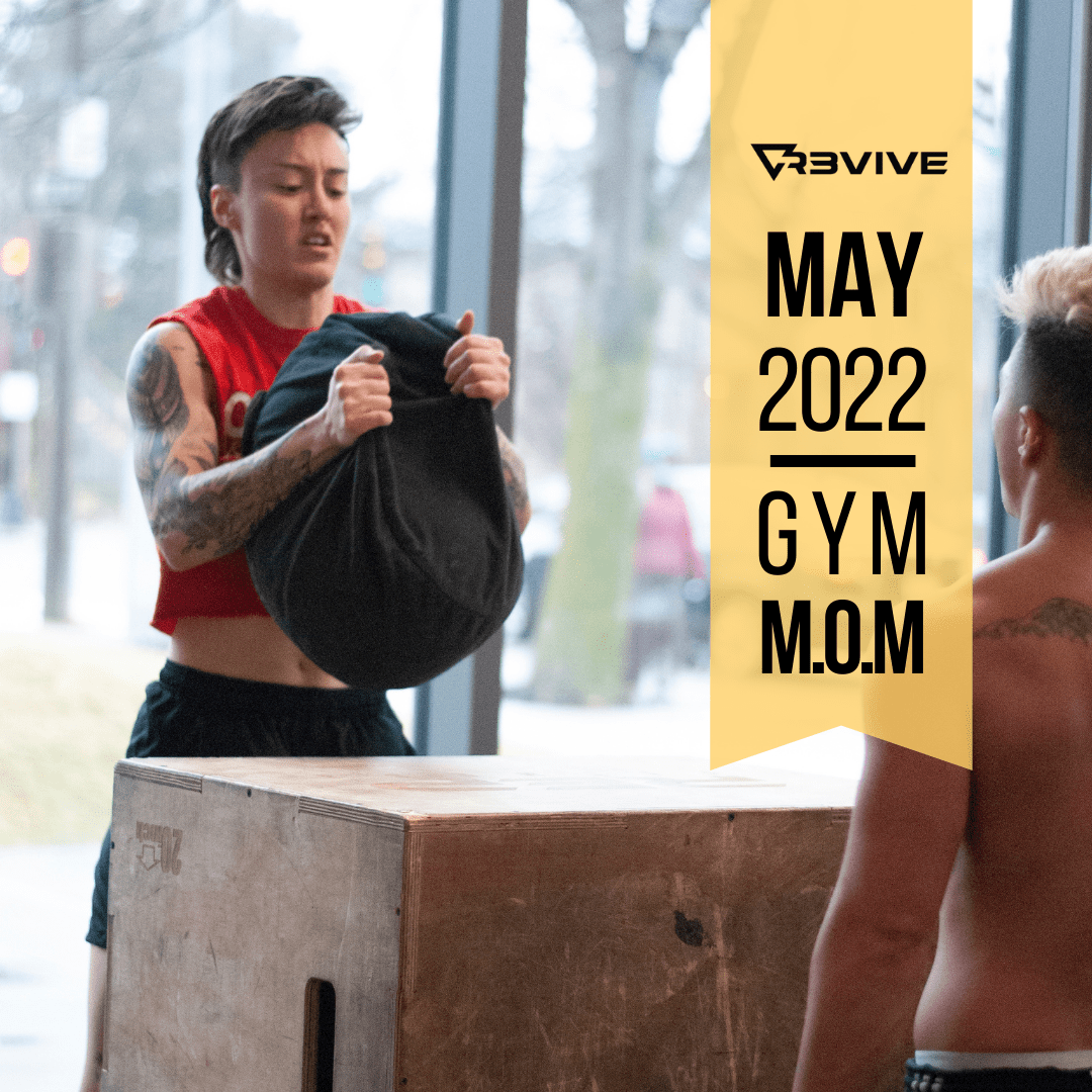 Woman lifting weighted bag onto wooden box, another person watches in gym; May 2022 Gym M.O.M promotion.