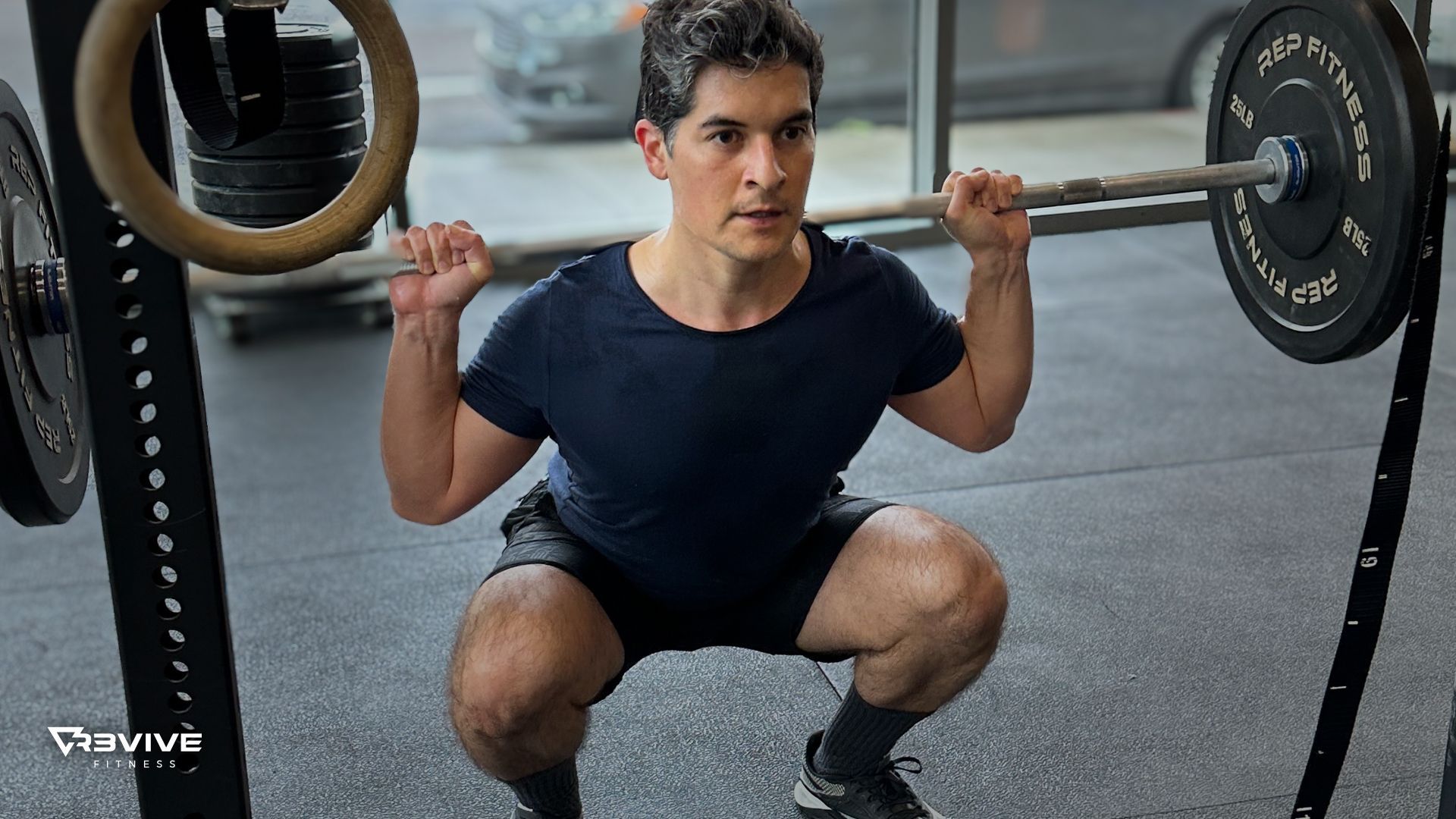 Man performing a barbell squat in a gym, wearing dark blue shirt and shorts.