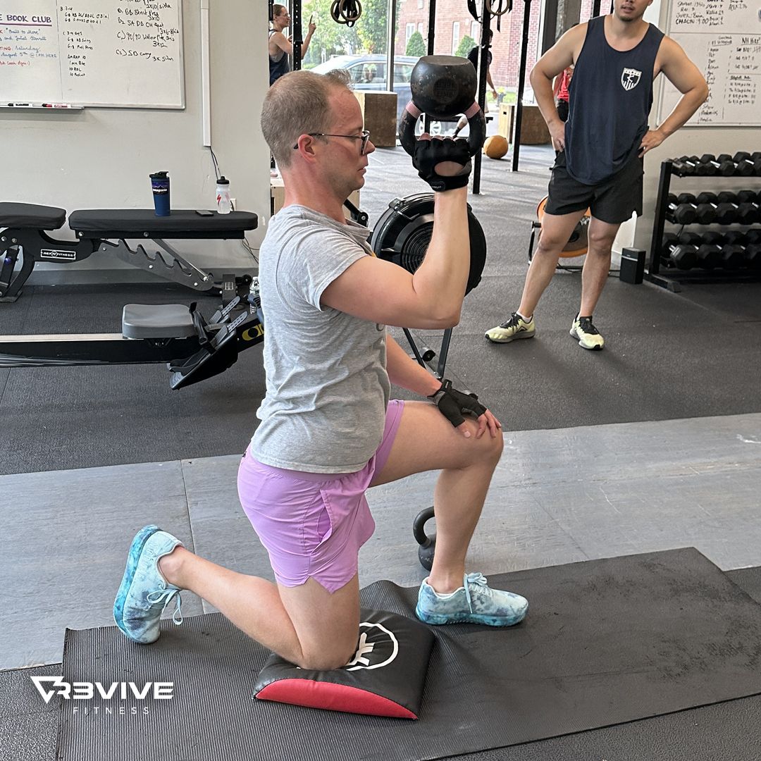 Man kneels, presses kettlebell overhead. Gym setting. Wearing grey shirt, pink shorts, holding a weight.