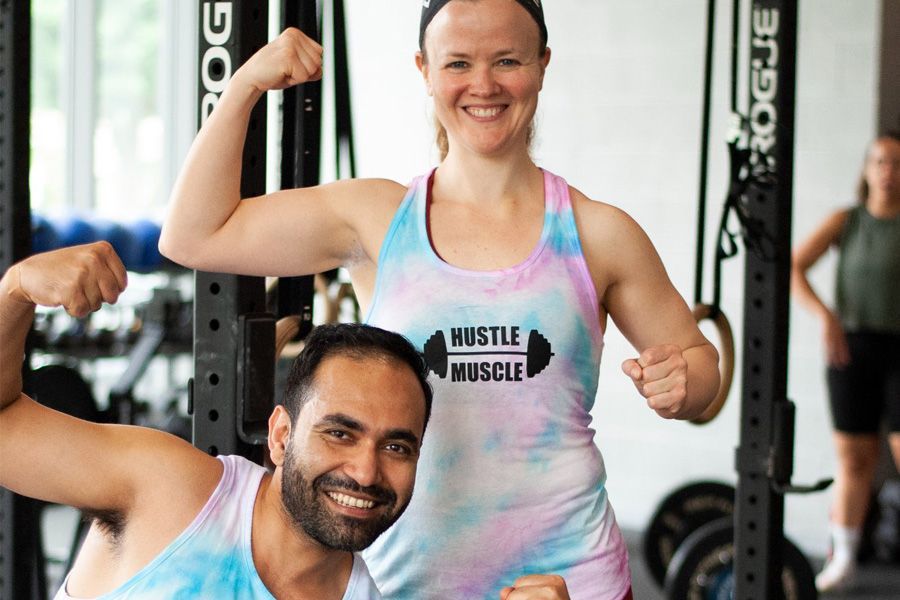 Two people flexing muscles in a gym. Woman in tie-dye tank top and man smiling.