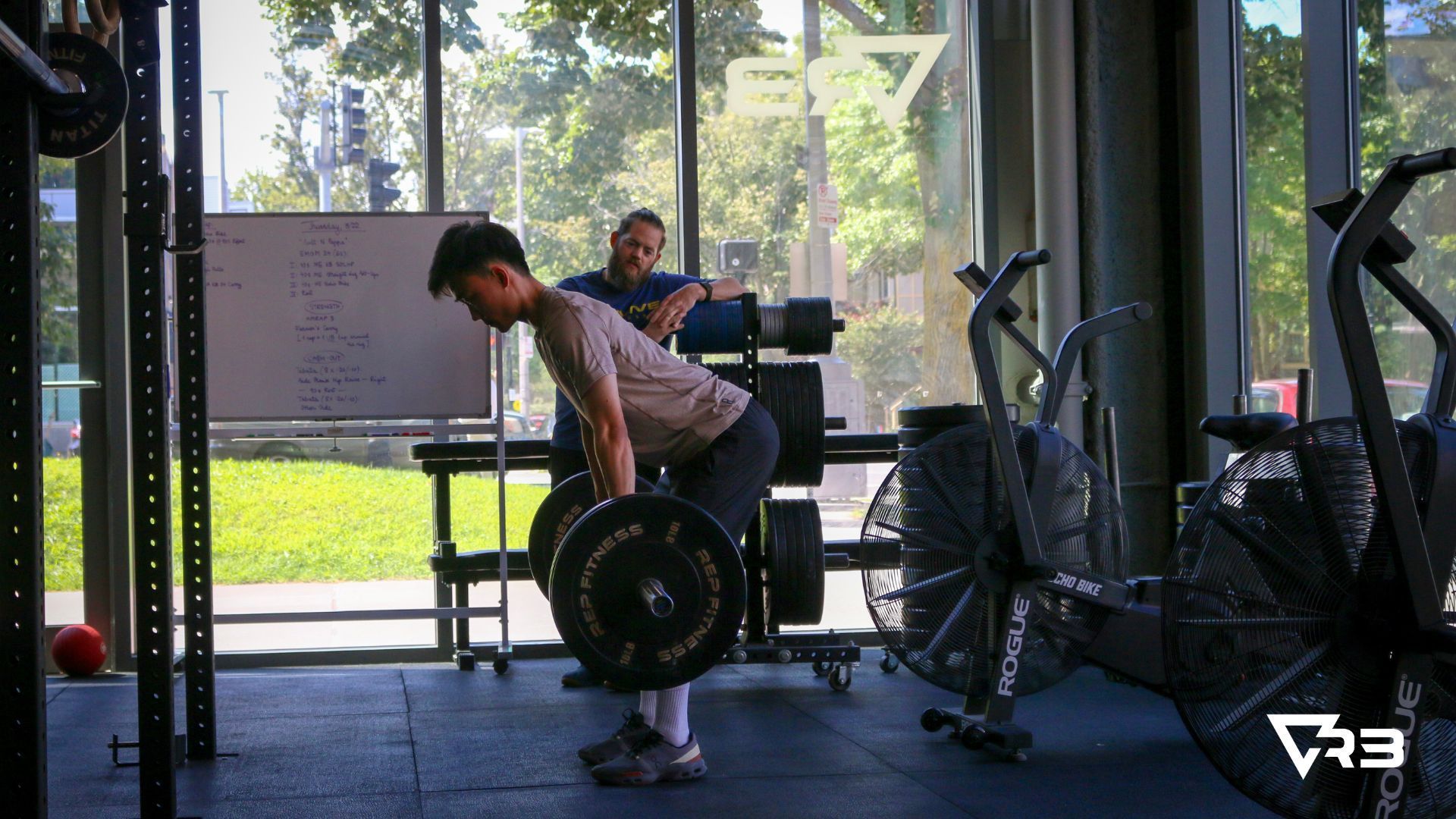 Man performing a barbell deadlift in a gym, with a trainer observing.