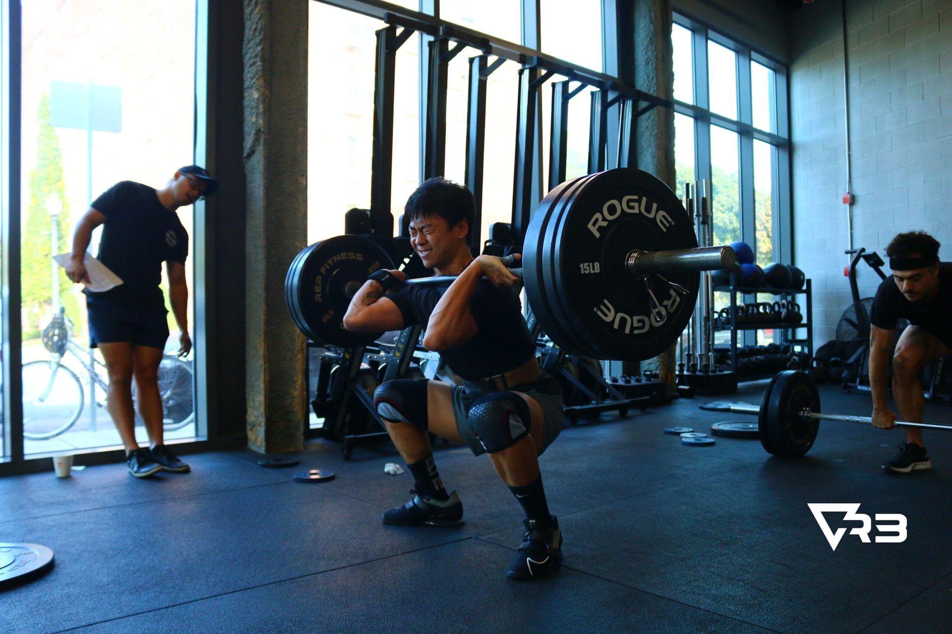 A man squats with a heavy barbell in a gym; two other men watch.