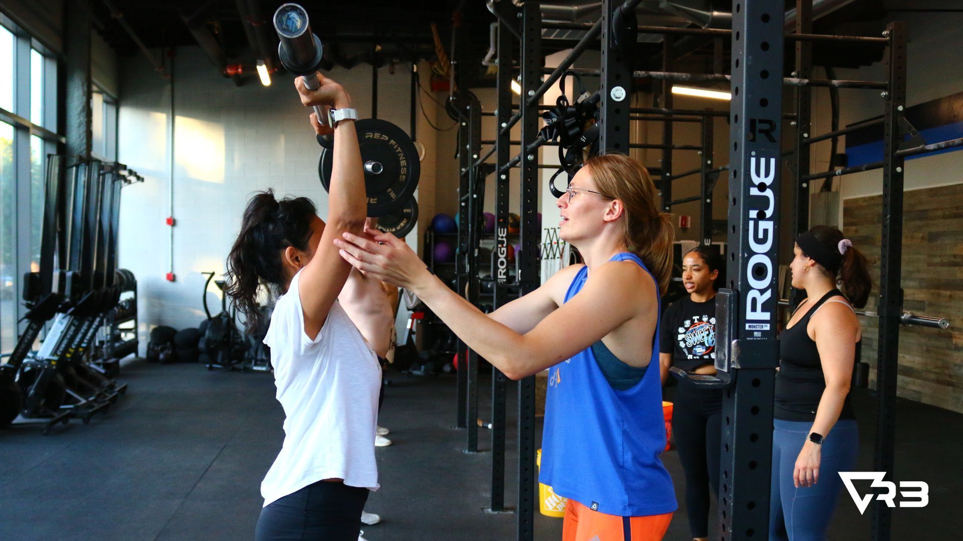 Woman lifting weight overhead, coached by another woman in a gym. Other people observe.