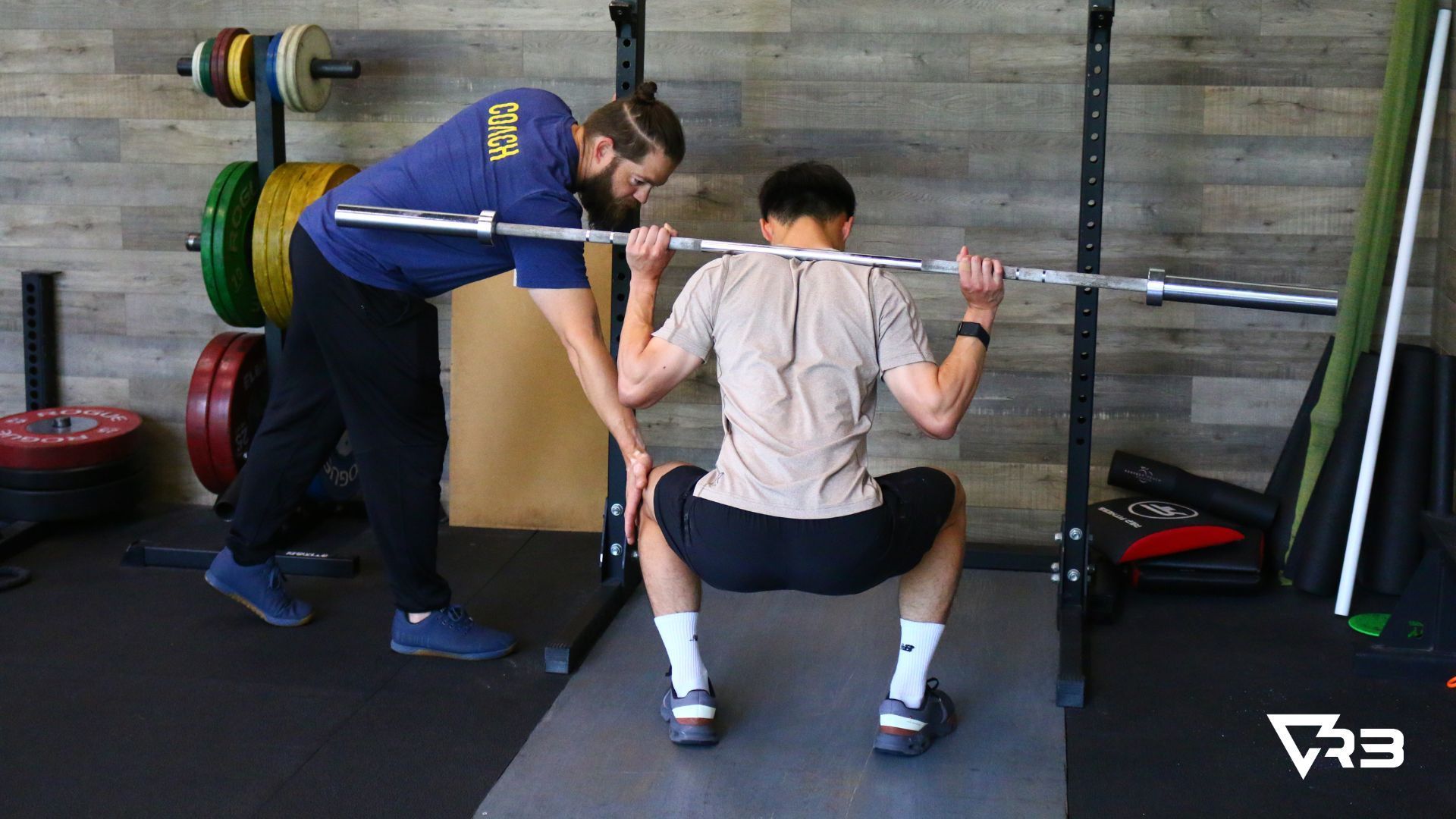 Man coaching another on barbell squat form. Gym setting; both men are focused.