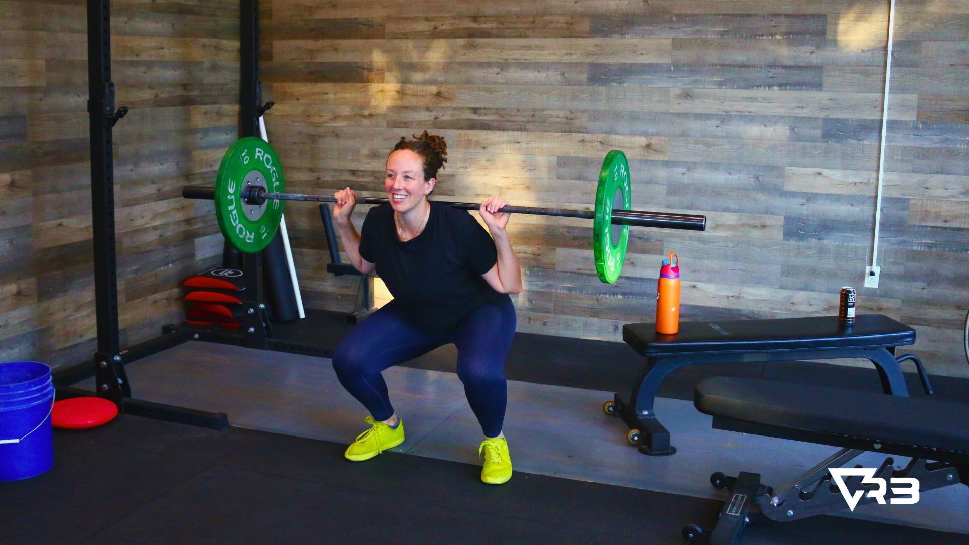 Woman smiles while squatting with barbell in a home gym; green and red weights, water bottle.