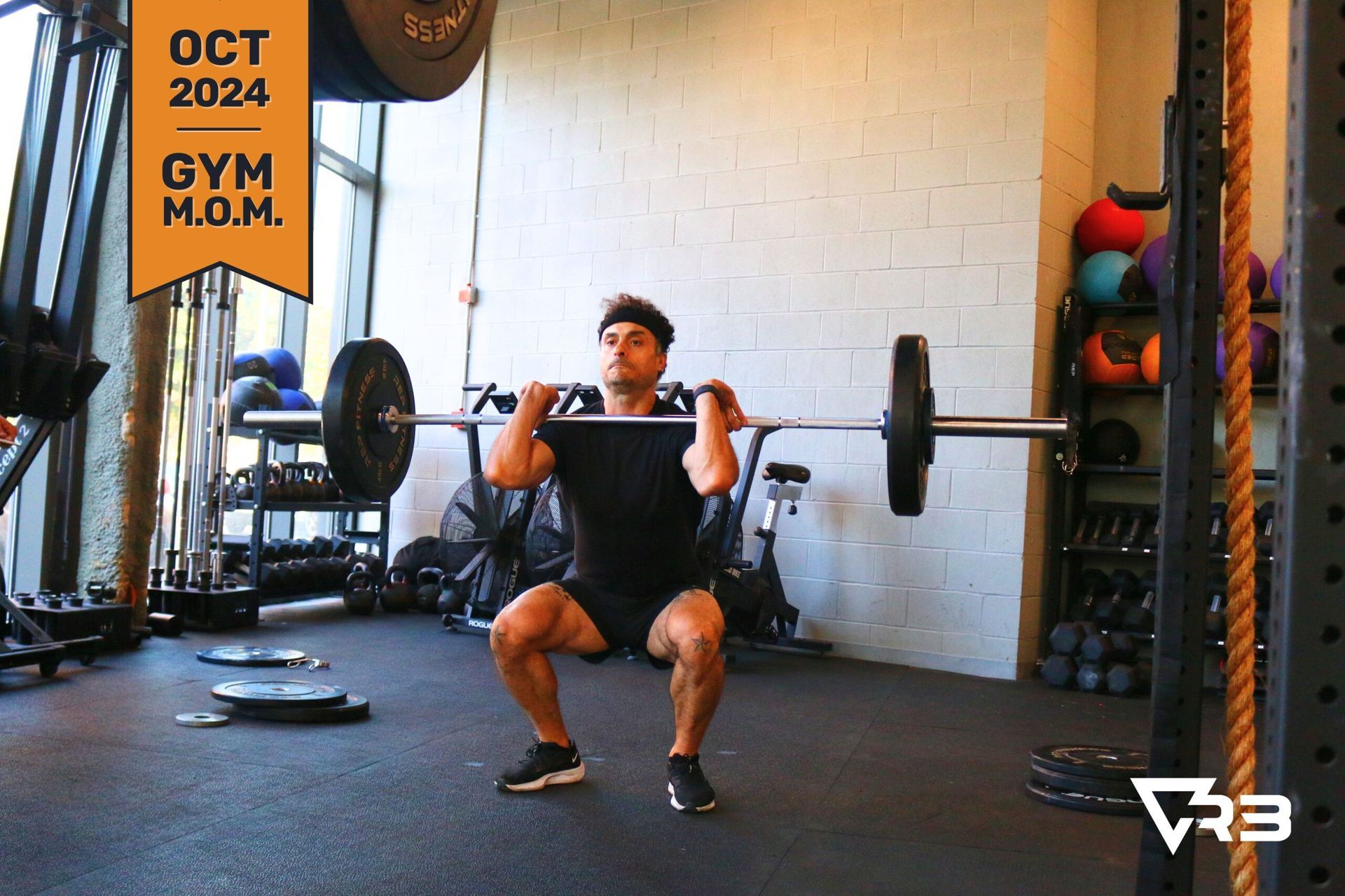Man in gym performs front squat with barbell. He is in a squat position, holding bar at shoulder level.