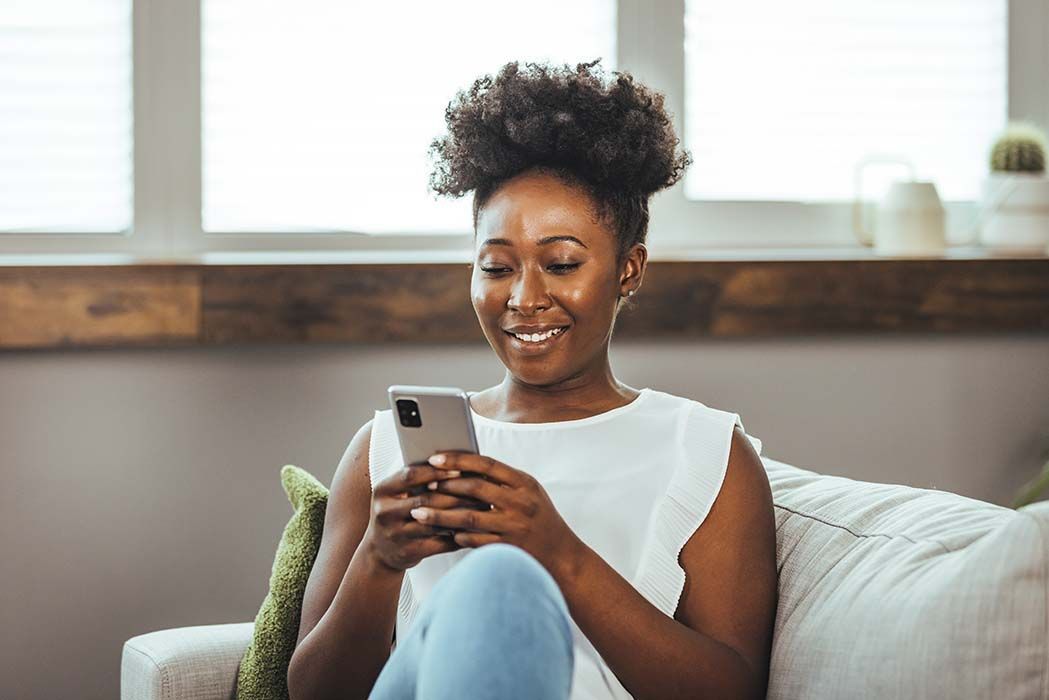 Smiling woman sitting on a couch, looking at her phone in a bright living room.
