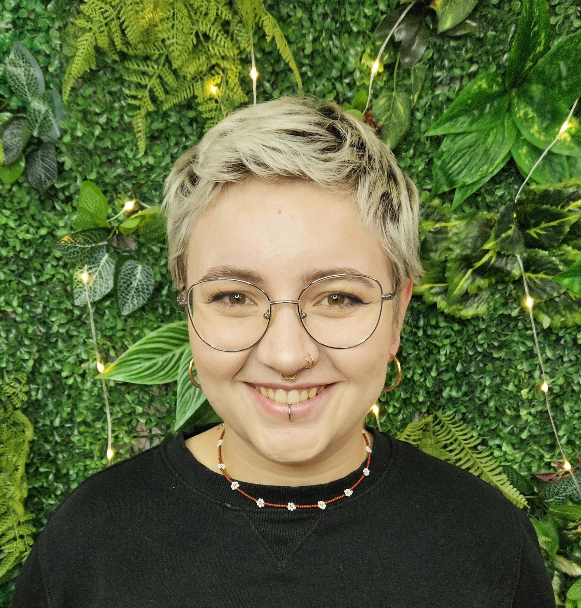 Eine Frau mit Brille und Nasenring lächelt vor einer grünen Wand.