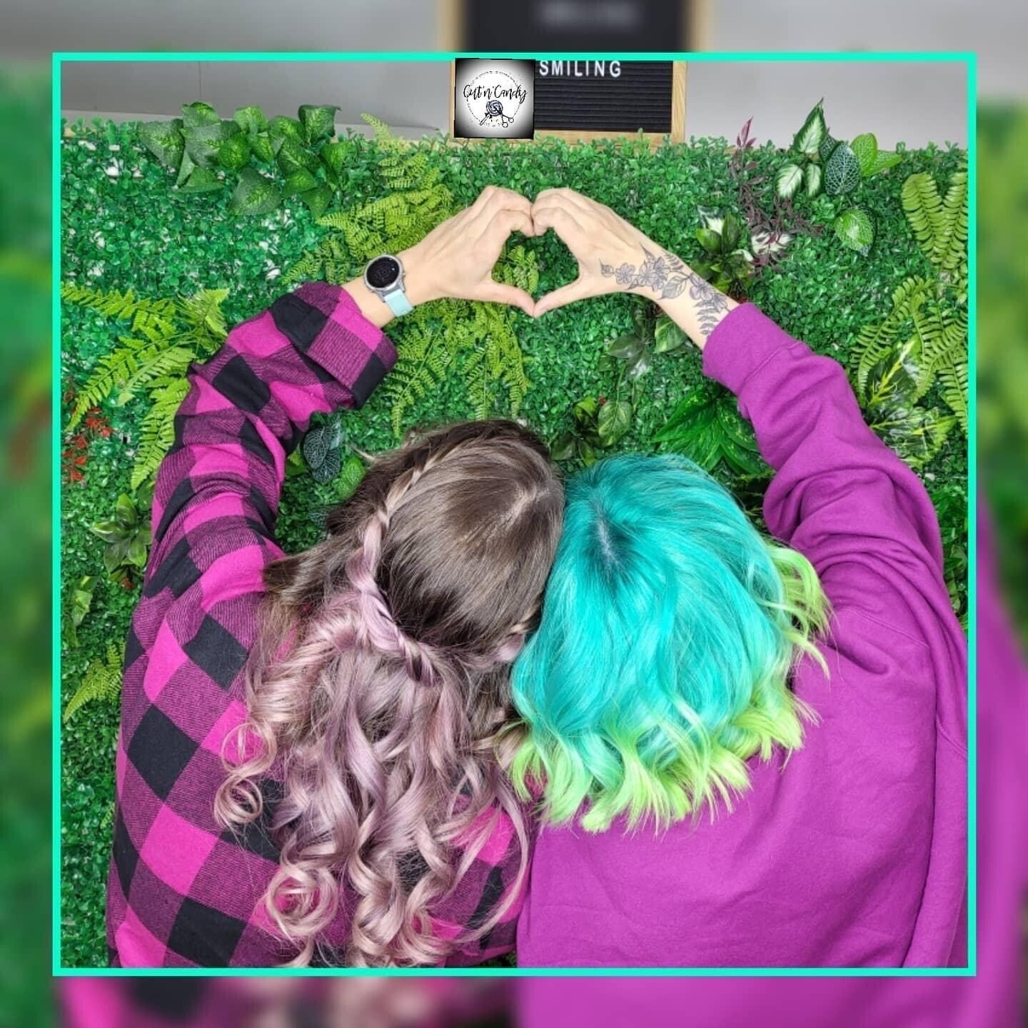 Zwei Frauen mit grünem und rosa Haar formen mit ihren Händen ein Herz.