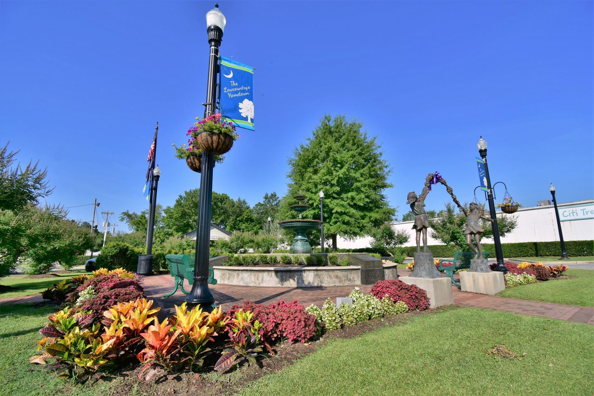 Park with a fountain, flower beds, sculptures, and lampposts under a blue sky.