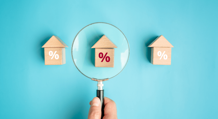 Hand holding magnifying glass over house-shaped blocks with percent signs, set against a blue backdrop.