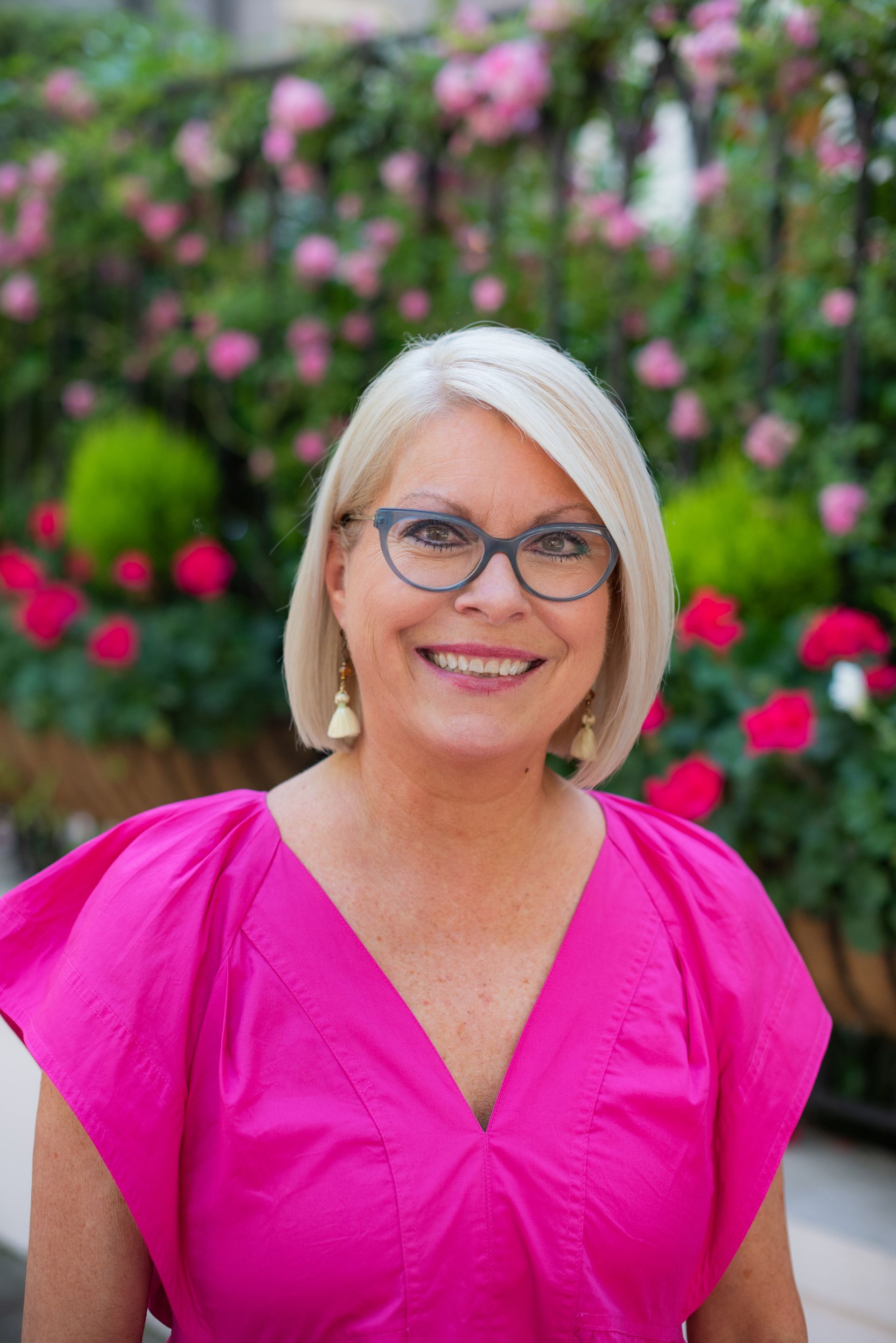 A woman wearing glasses and a pink top is smiling in front of flowers.