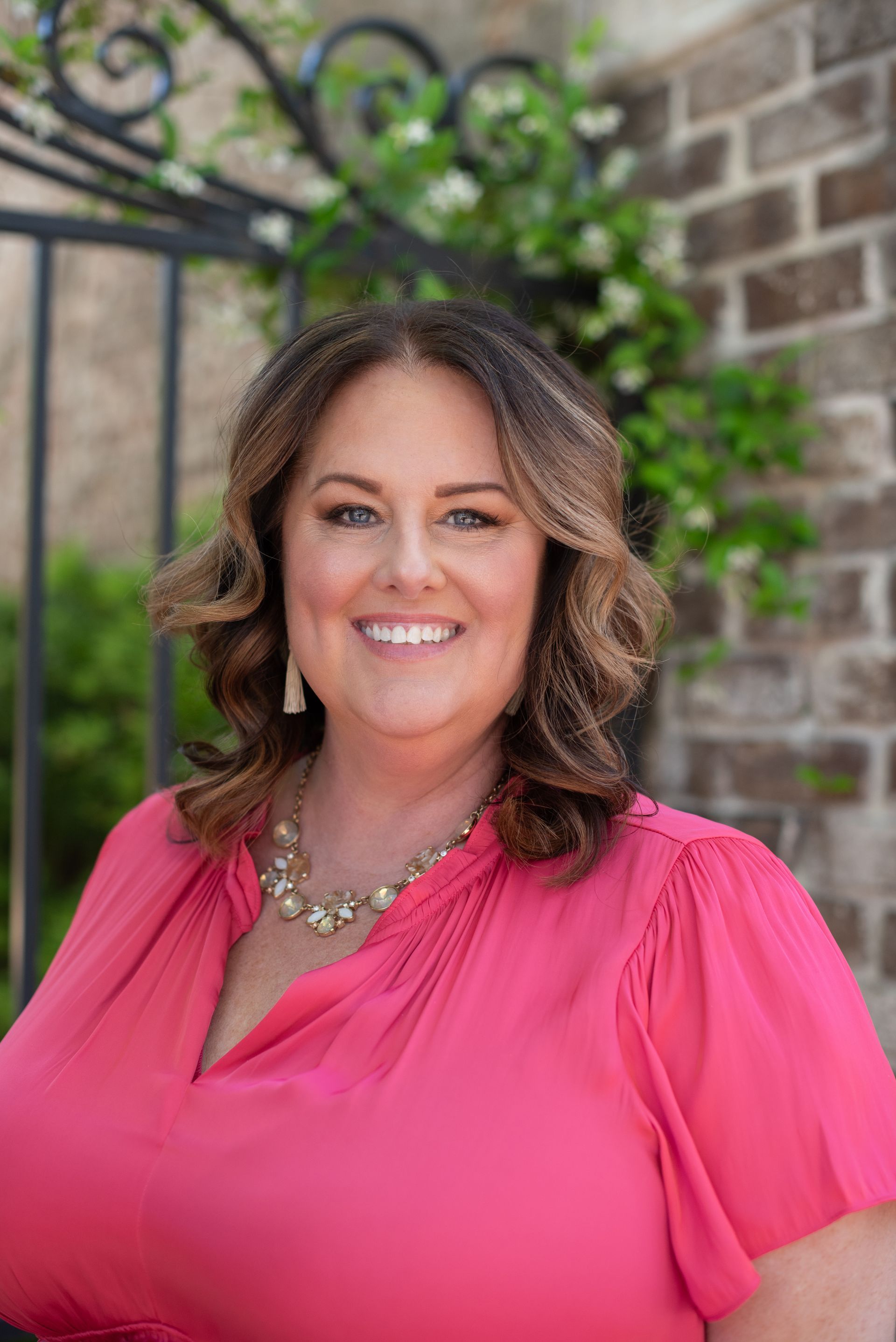 A woman in a pink shirt and necklace is smiling in front of a brick wall.