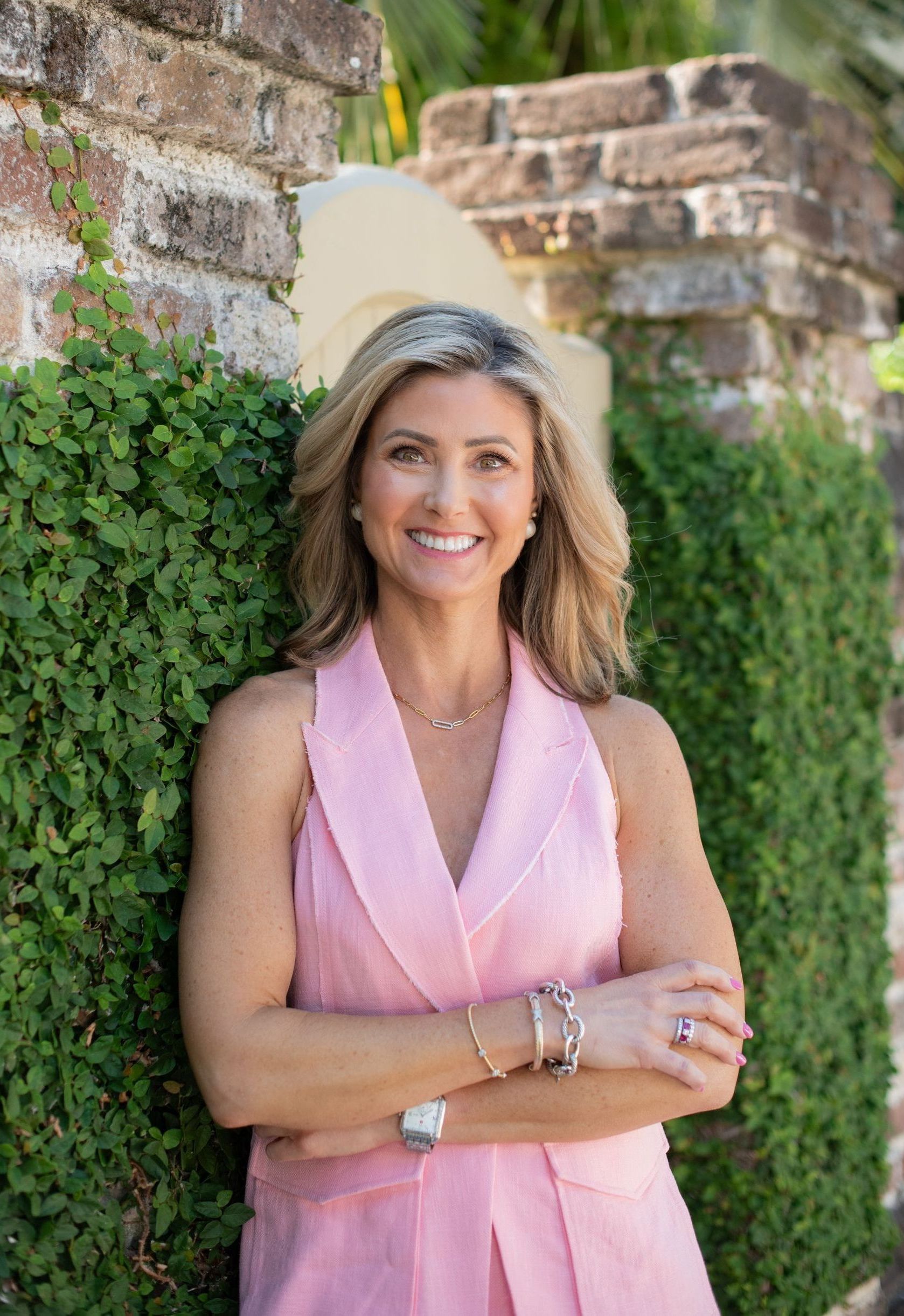 A woman in a pink dress is standing in front of a brick wall with her arms crossed.