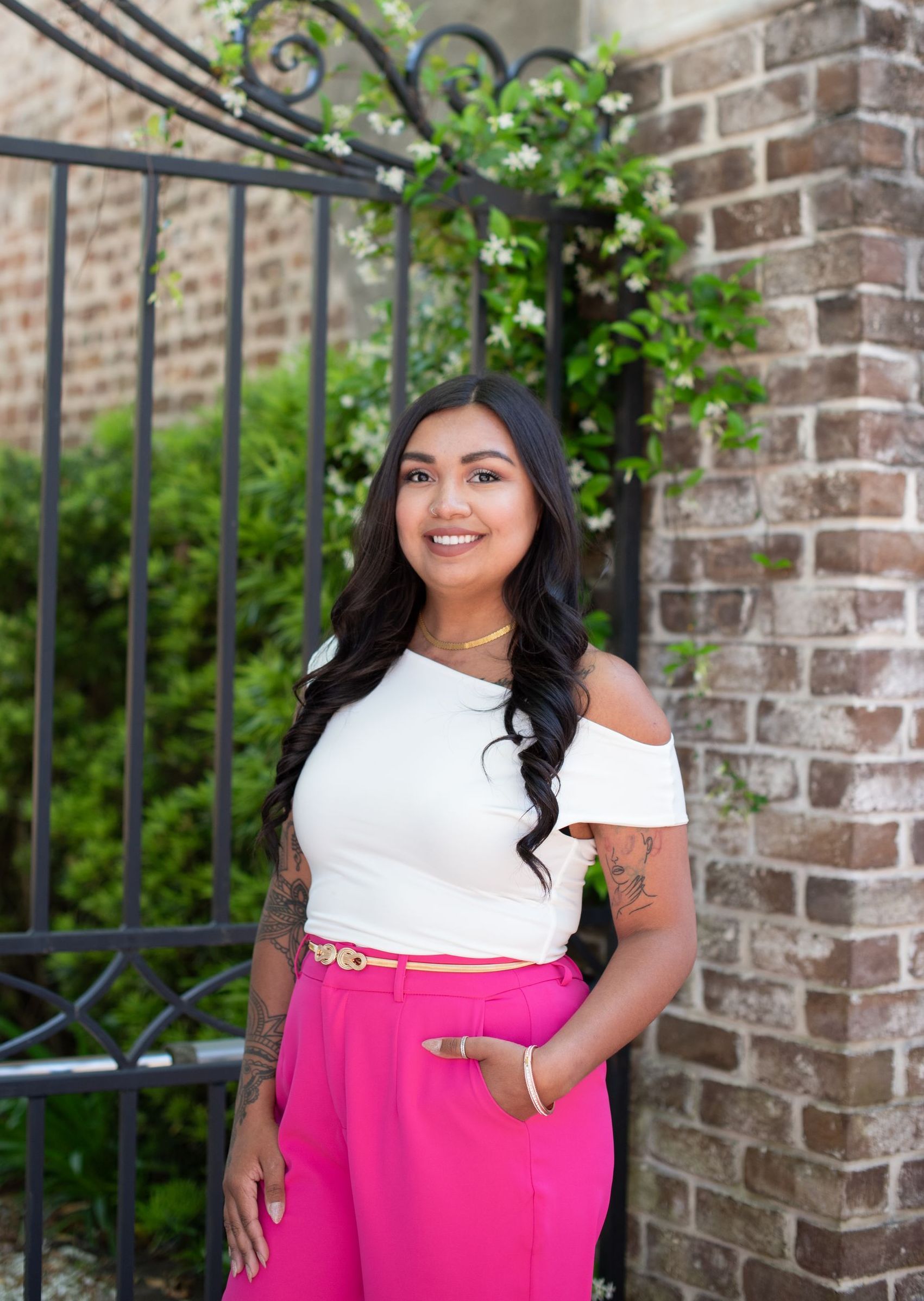 A woman in a white top and pink pants is standing in front of a brick wall.