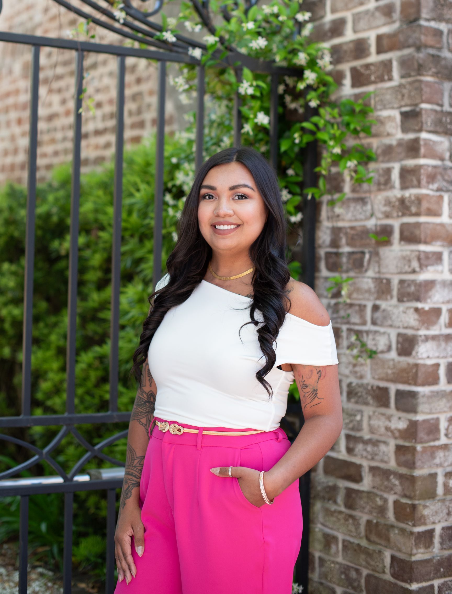 A woman in a white off the shoulder top and pink pants is standing in front of a brick wall.