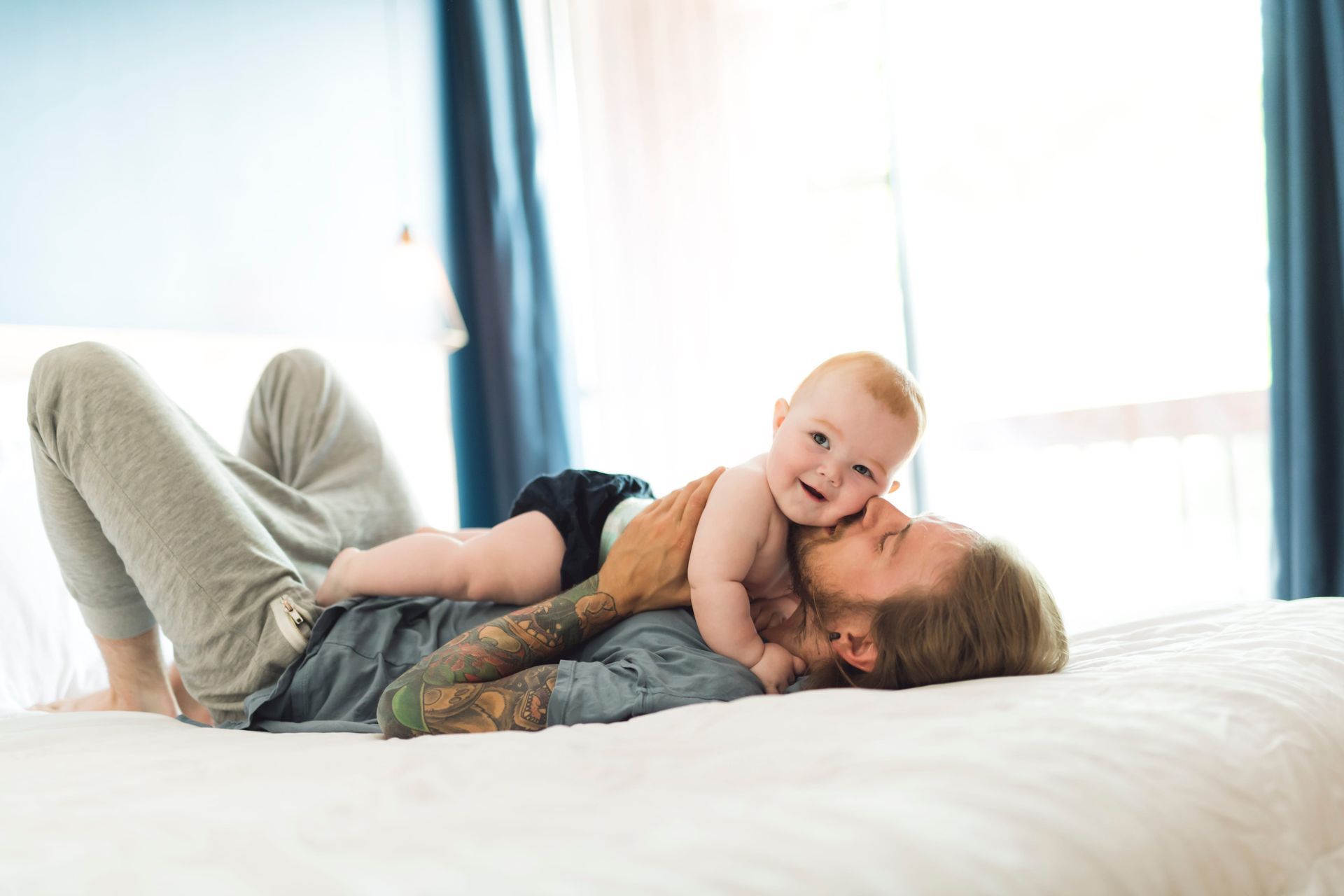 Adult lying on a bed with a smiling baby on top, in a bright bedroom