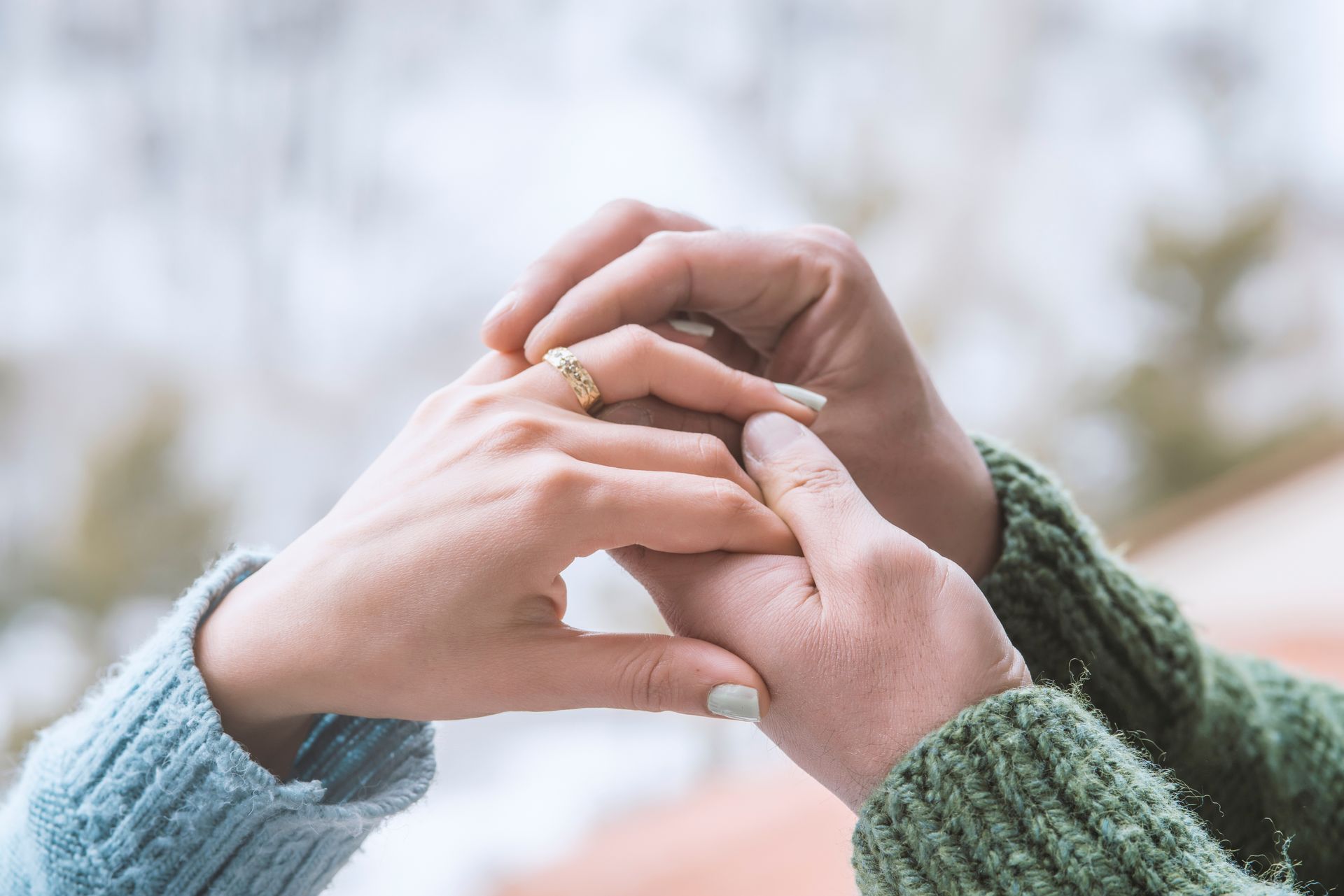 Hands exchanging a ring; one hand wearing a gold ring with a gemstone, both in sweaters with a blurred background.