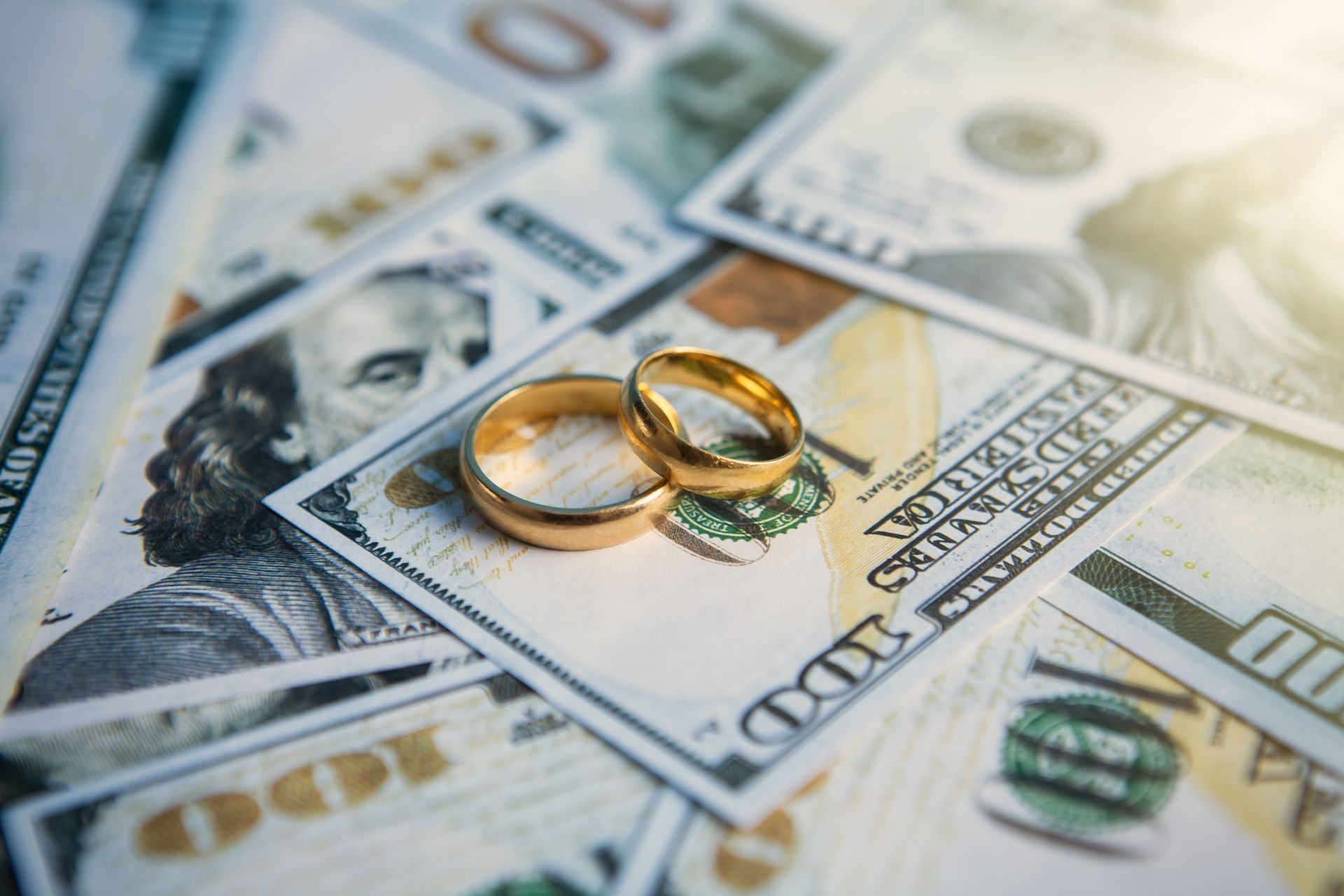 Two gold wedding rings on top of scattered hundred dollar bills.