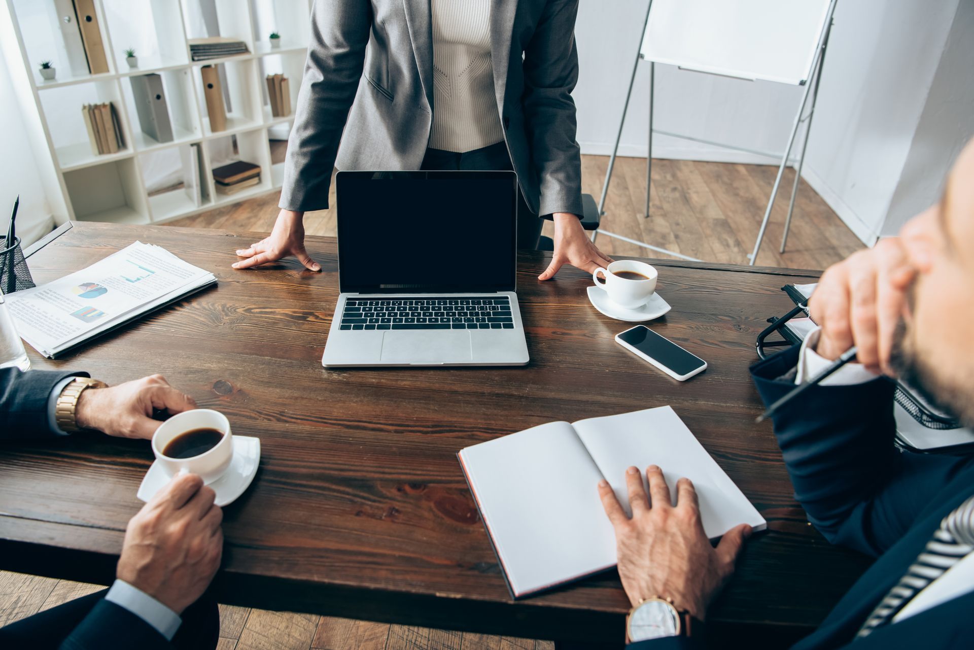 Business meeting at a wooden table. A woman stands, others sit with coffee, laptop, and notebooks.