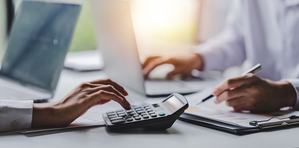Hands using a calculator and writing on documents with a pen at a desk with a laptop.
