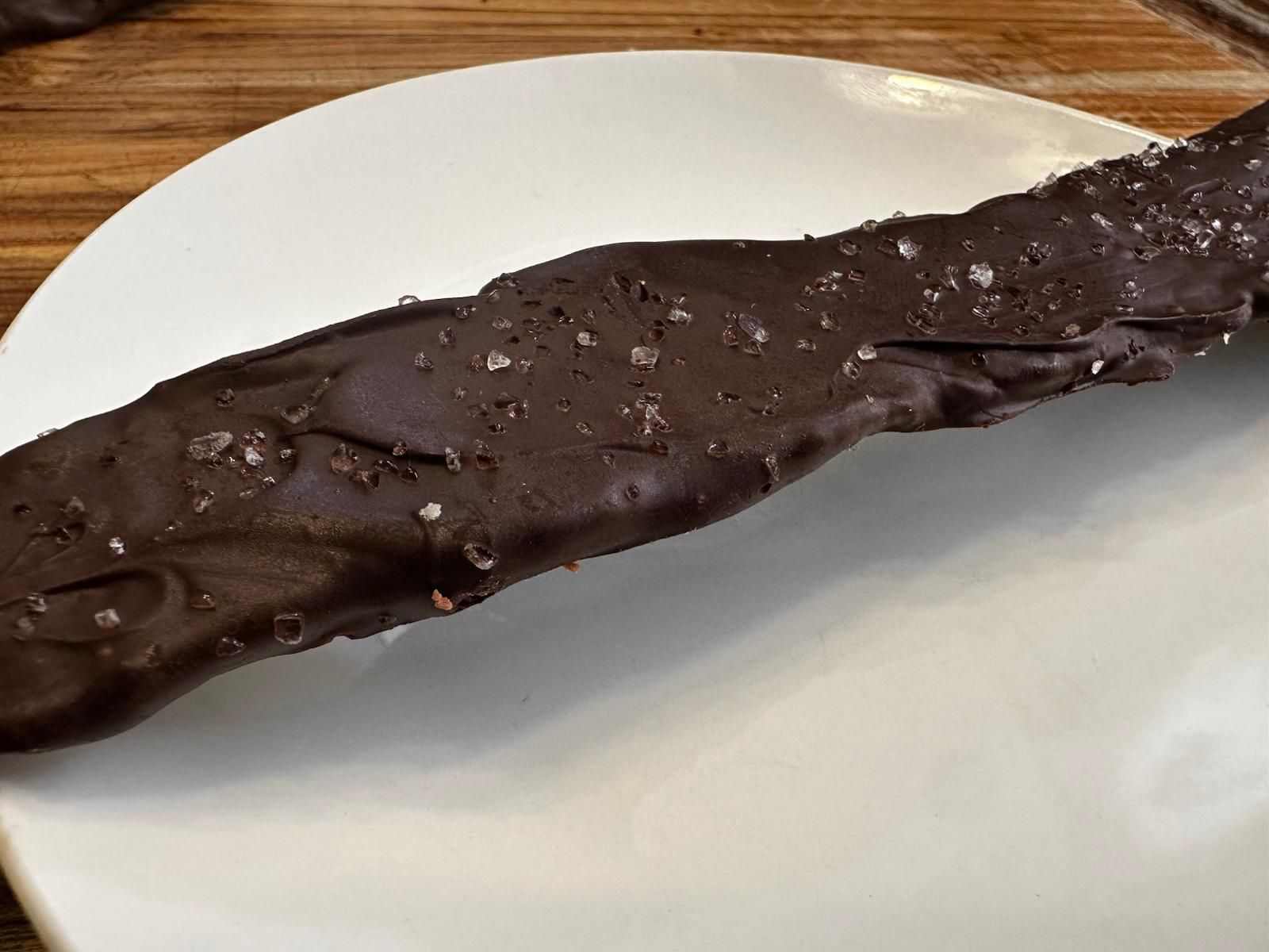 Dark chocolate-covered pretzel rod on a white plate, sprinkled with coarse salt.