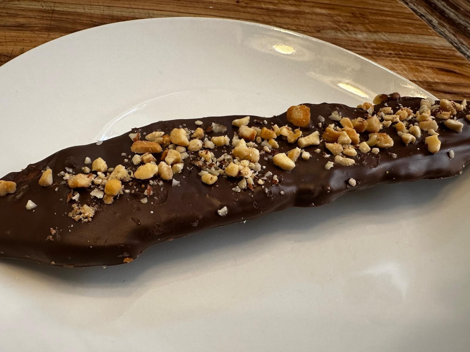 Chocolate-covered banana topped with chopped peanuts on a white plate.