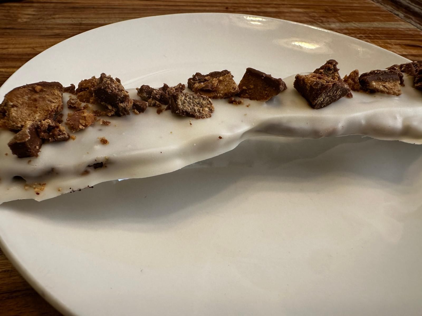 White frosted dessert topped with crumbled chocolate pieces on a white plate, resting on a wooden table.